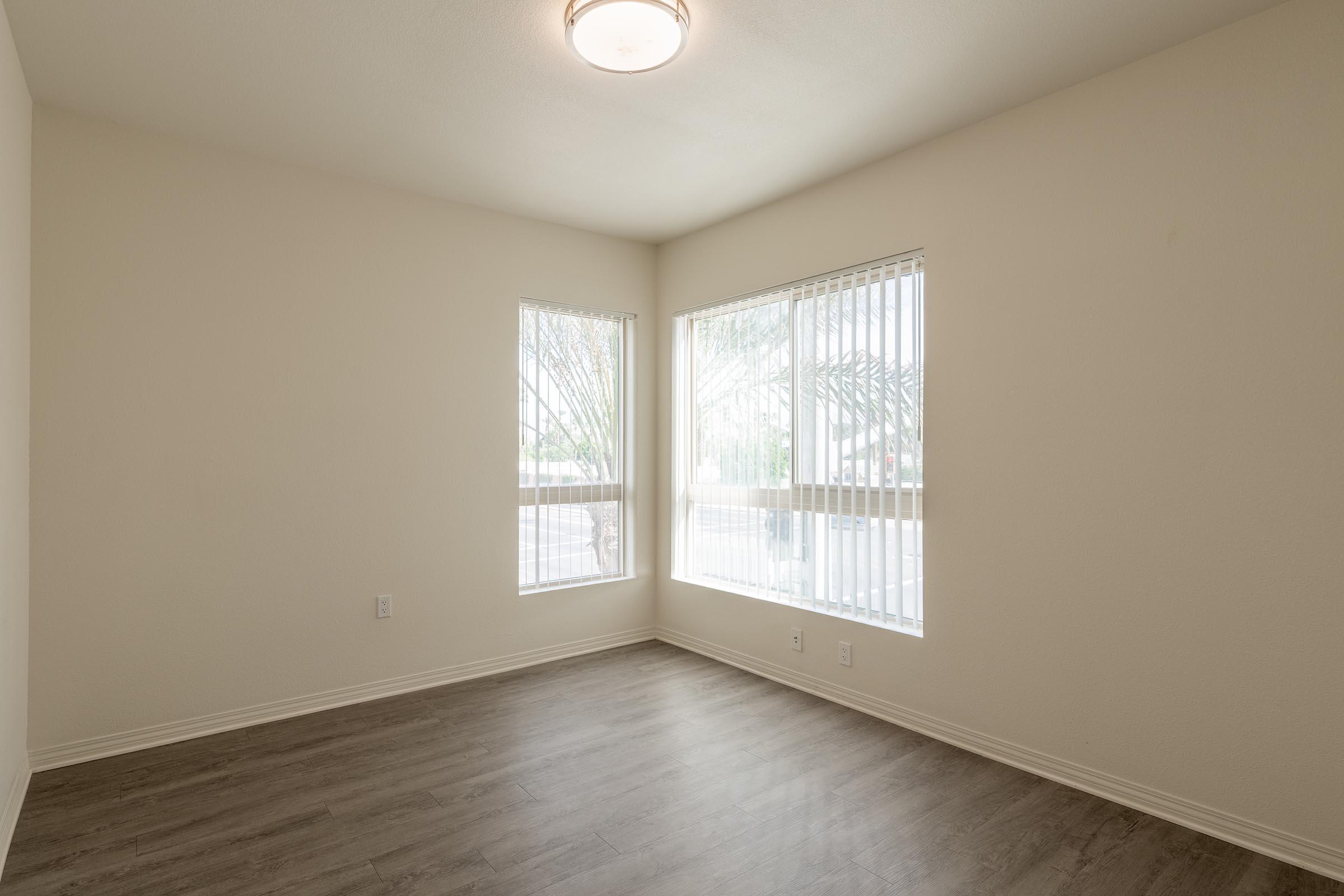 A well-lit empty room with two large windows featuring vertical blinds, showcasing a wooden floor. The walls are light-colored, creating a spacious feel, and a ceiling light fixture is visible.
