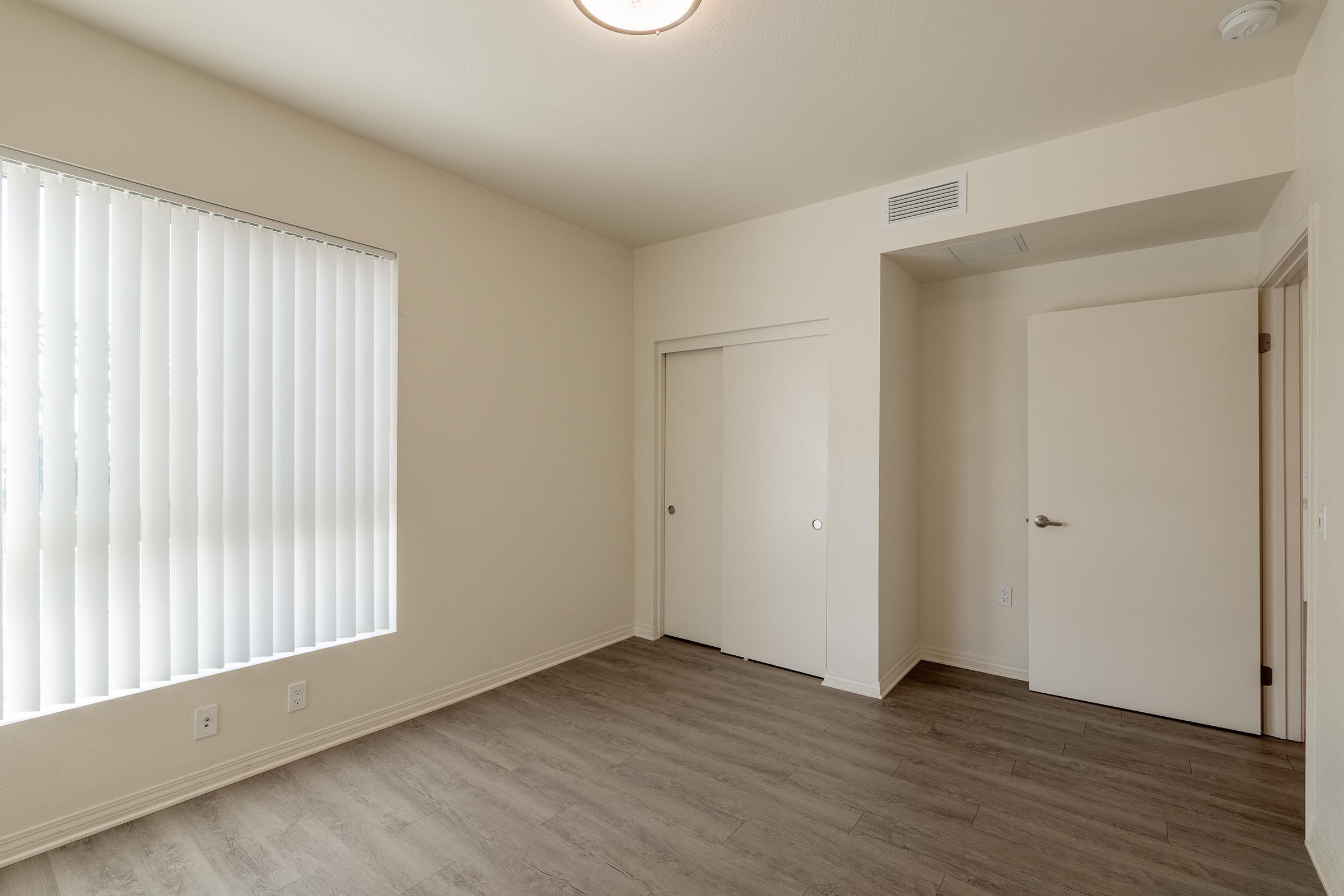 Empty room with light-colored walls and laminate flooring. One wall features vertical blinds over a window, allowing natural light. A closet with double doors is situated along the adjacent wall, and a plain door leads to another area. The ceiling light provides illumination.
