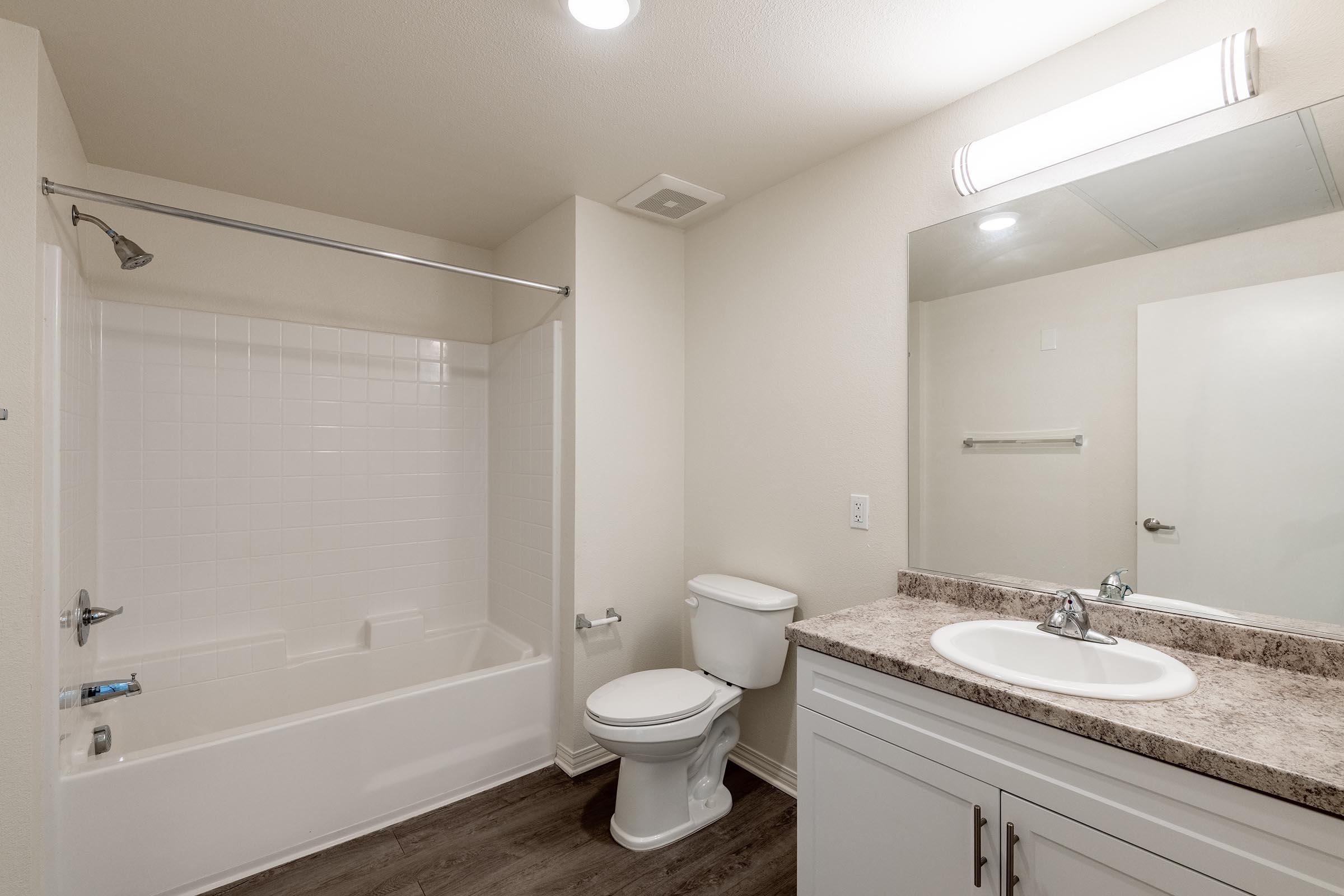 A bathroom featuring a white bathtub with a shower, a modern toilet, and a sink with a countertop. The walls are light-colored, and there is a large mirror above the sink. The flooring is a wood-like laminate. The overall design is clean and contemporary.