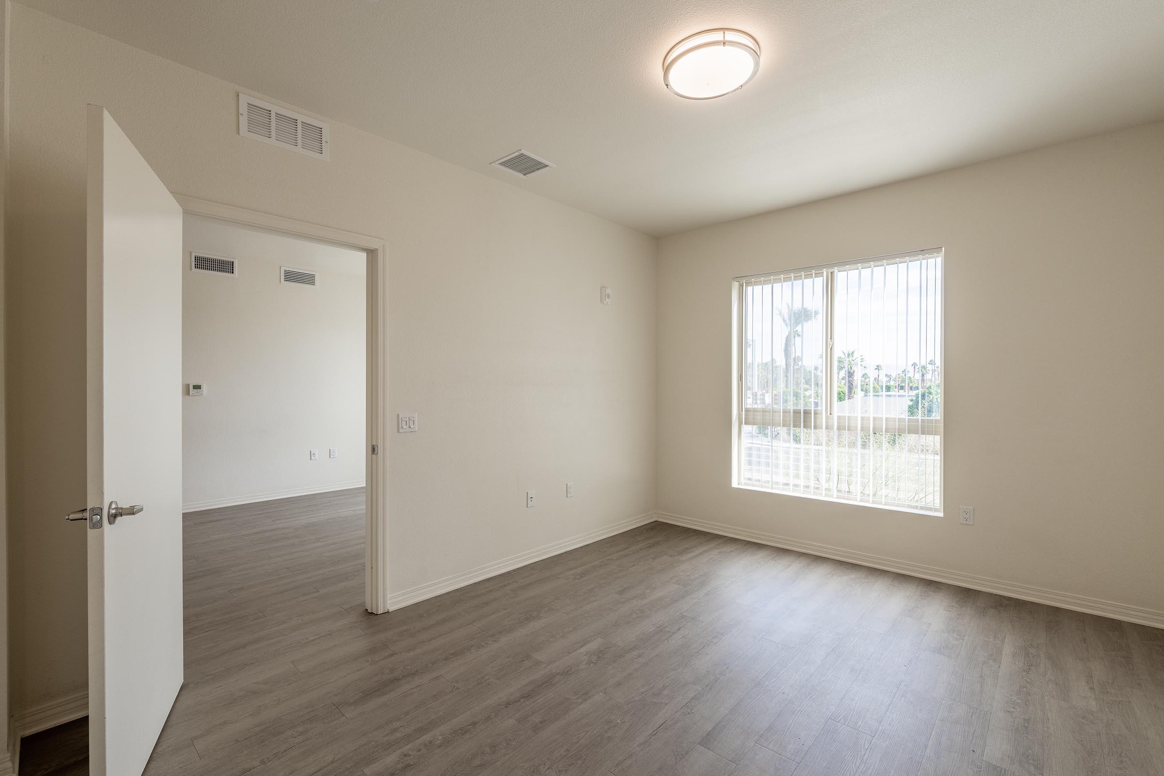 A partially furnished room with light-colored walls and wooden laminate flooring. A door leads to another room in the background. The window allows natural light to enter, illuminating the space, which appears clean and spacious. A ceiling light fixture provides additional illumination.