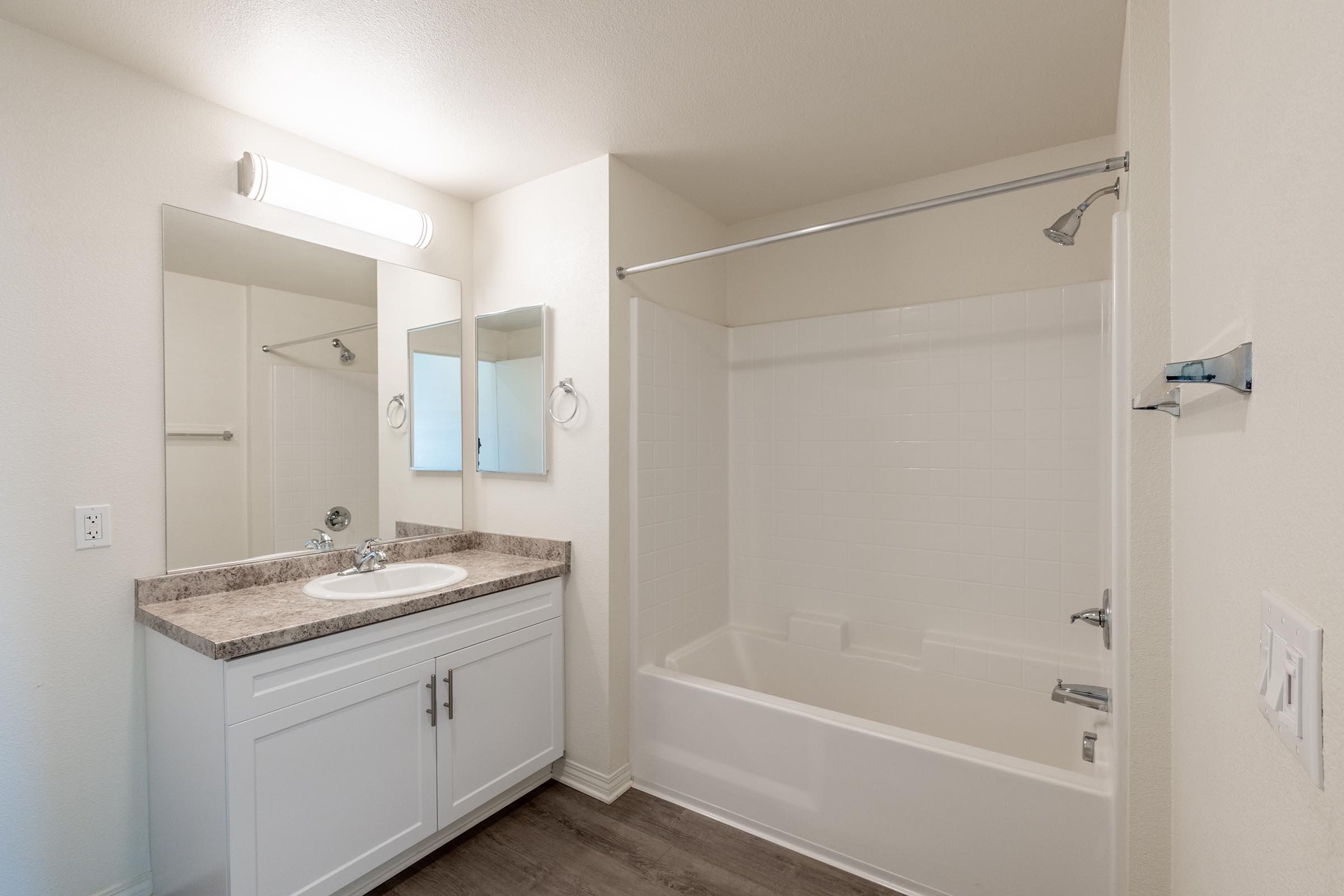 A well-lit bathroom featuring a white bathtub with a showerhead, a single sink with a mirrored cabinet above, and light-colored walls. The flooring is a dark wood laminate, and there are two mirrors mounted on the wall, creating a spacious feel. The overall design is modern and minimalistic.