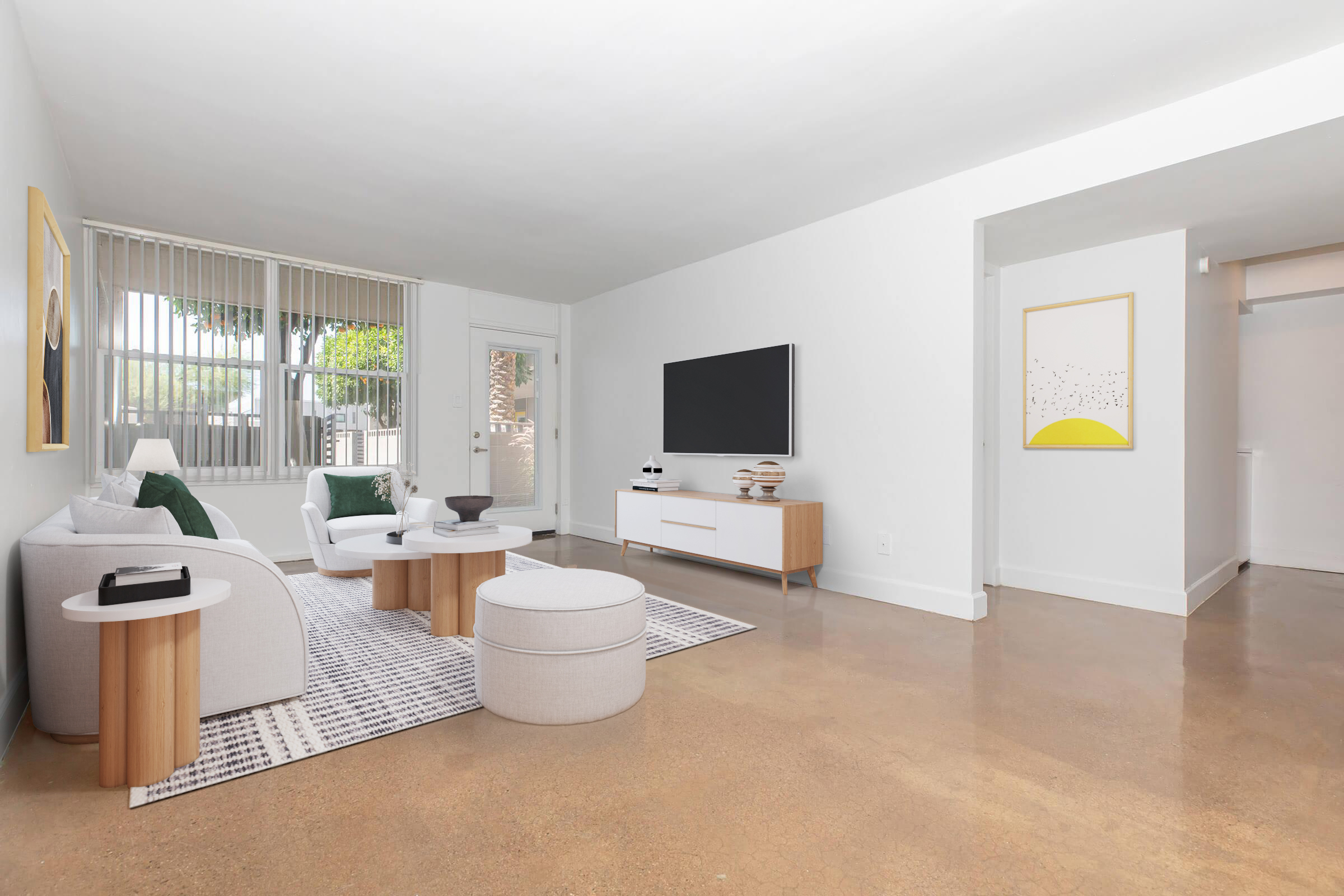 A modern, minimalist living room featuring a white sofa with gray accent pillows, a round ottoman, and a coffee table. The walls are painted white, with large windows allowing natural light. A flat-screen TV is mounted on the wall, and a framed abstract art piece hangs nearby. The floor has a polished concrete finish.