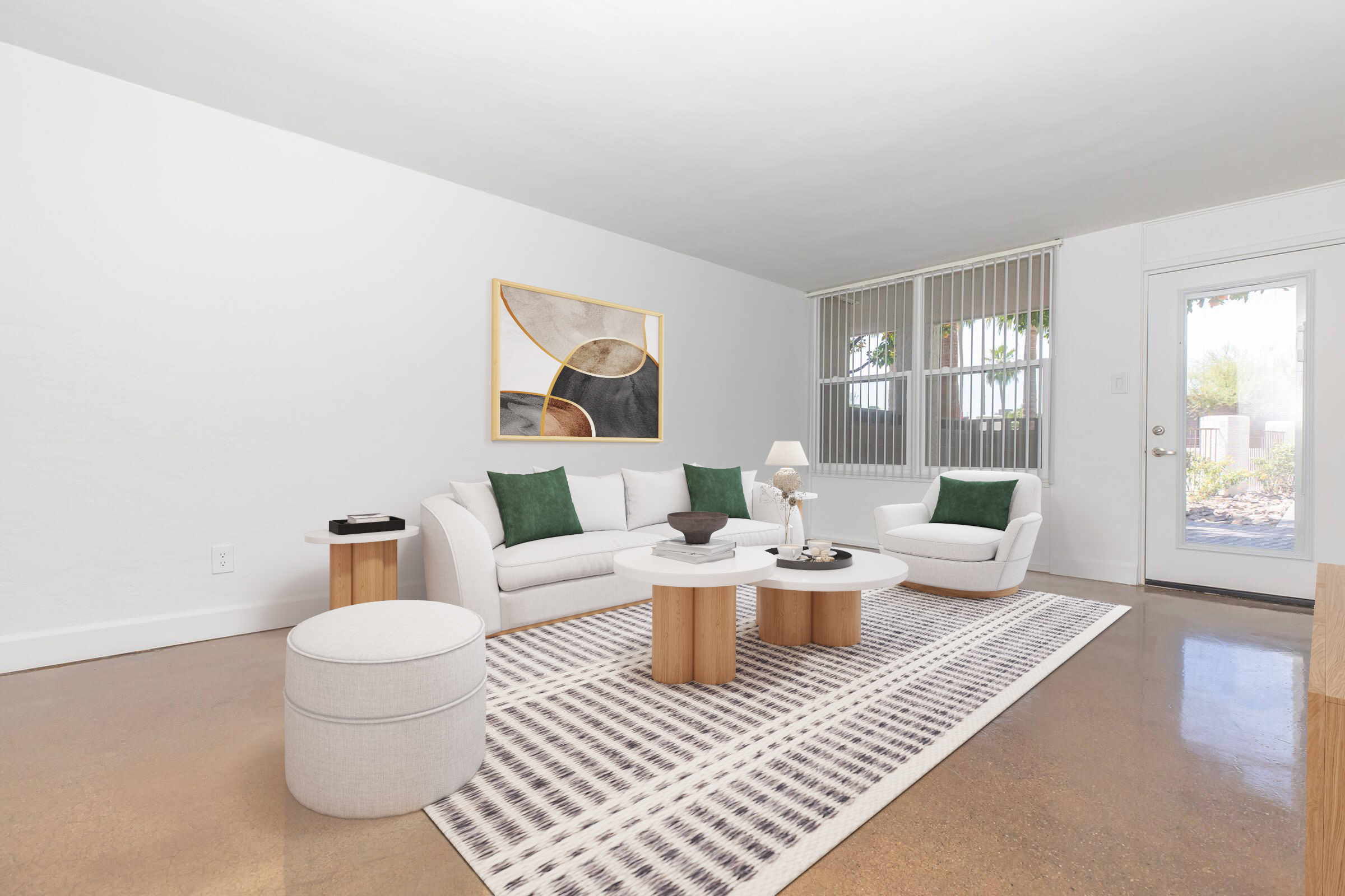 A modern living room featuring a light-colored sofa with green pillows, a round coffee table with decorative items, and a matching ottoman. The floor has a polished concrete finish, and a large area rug adds texture. Natural light filters in through a glass door, and abstract art hangs on the wall.