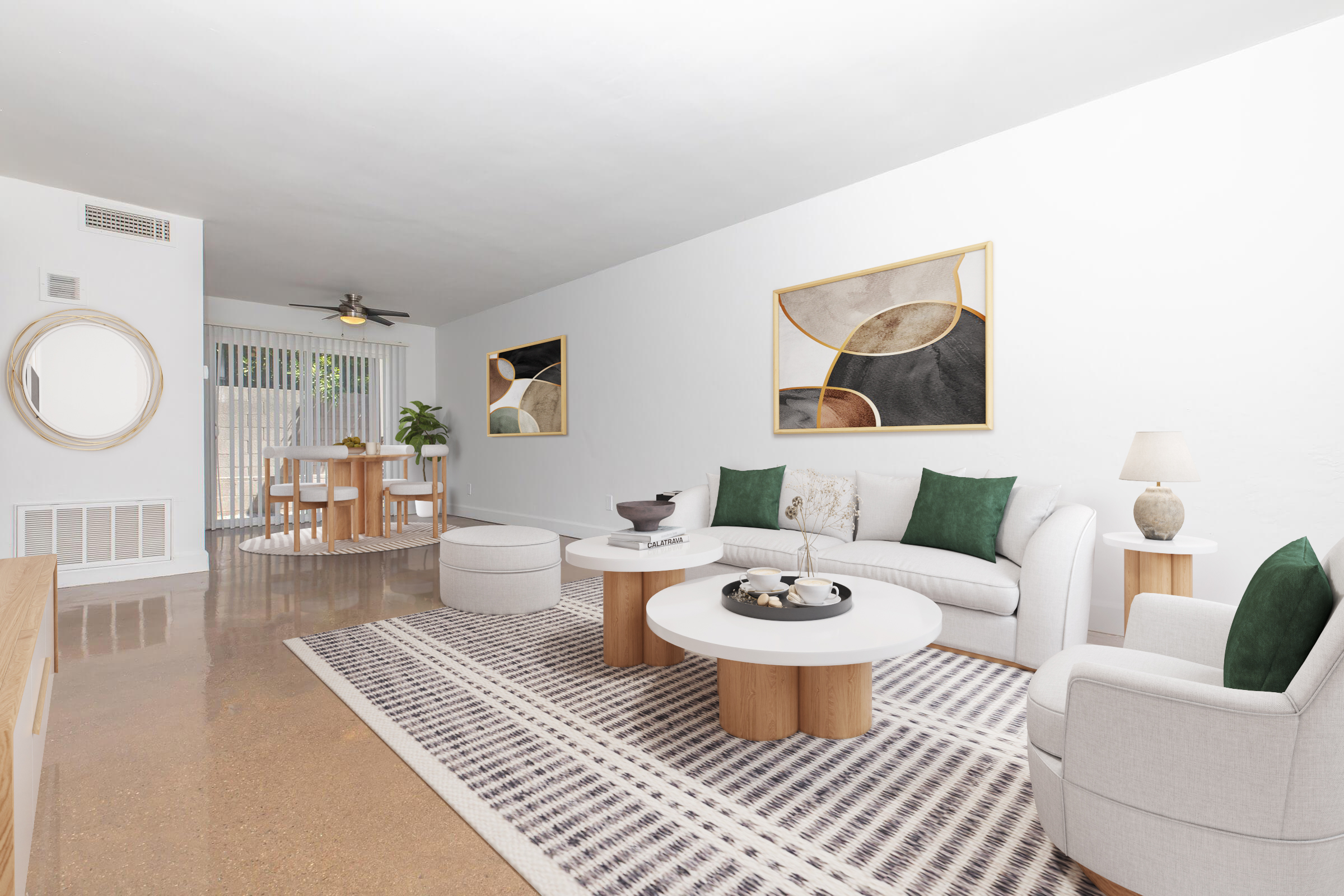 A modern living room featuring a light-colored sofa and lounge chairs arranged around circular wooden coffee and side tables. The space includes a geometric area rug, two large framed abstract art pieces on the wall, and a dining area with a round table in the background. Natural light streams in from a sliding door.