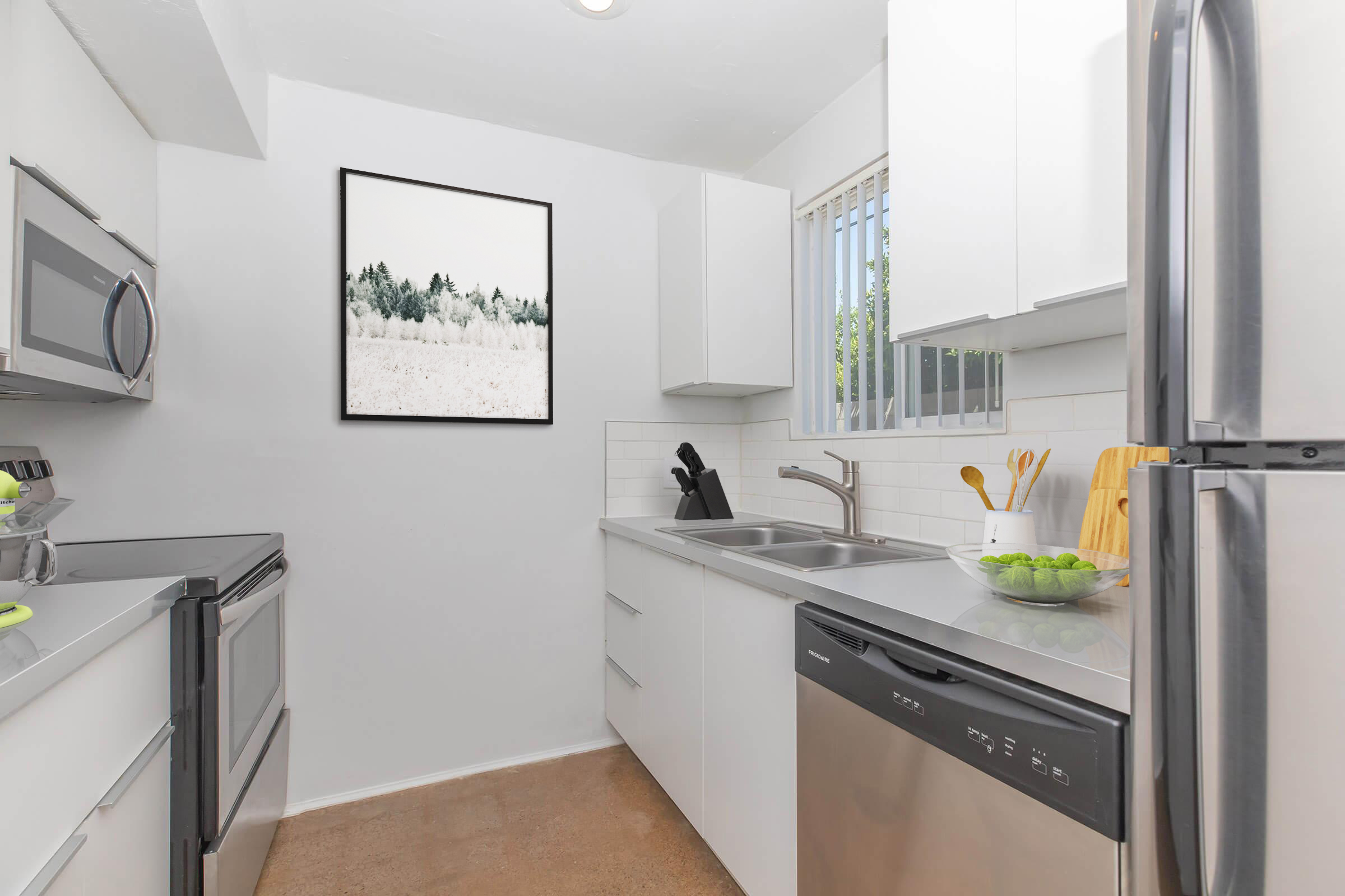 Modern kitchen featuring stainless steel appliances, including a microwave and refrigerator. The space has sleek white cabinets, a stainless steel sink, and a dishwasher. A framed black-and-white landscape photograph hangs on the wall, and a small bowl of green fruit is on the countertop, adding a fresh touch to the minimalist design.