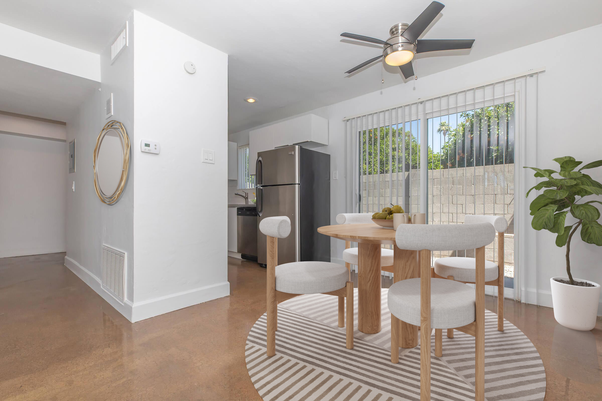 A bright dining area with a round wooden table and four light-colored chairs. A ceiling fan hangs above, and there are large sliding glass doors leading to an outdoor space. A potted plant adds greenery, and a sleek refrigerator is visible in the background, along with a modern, open-concept layout.