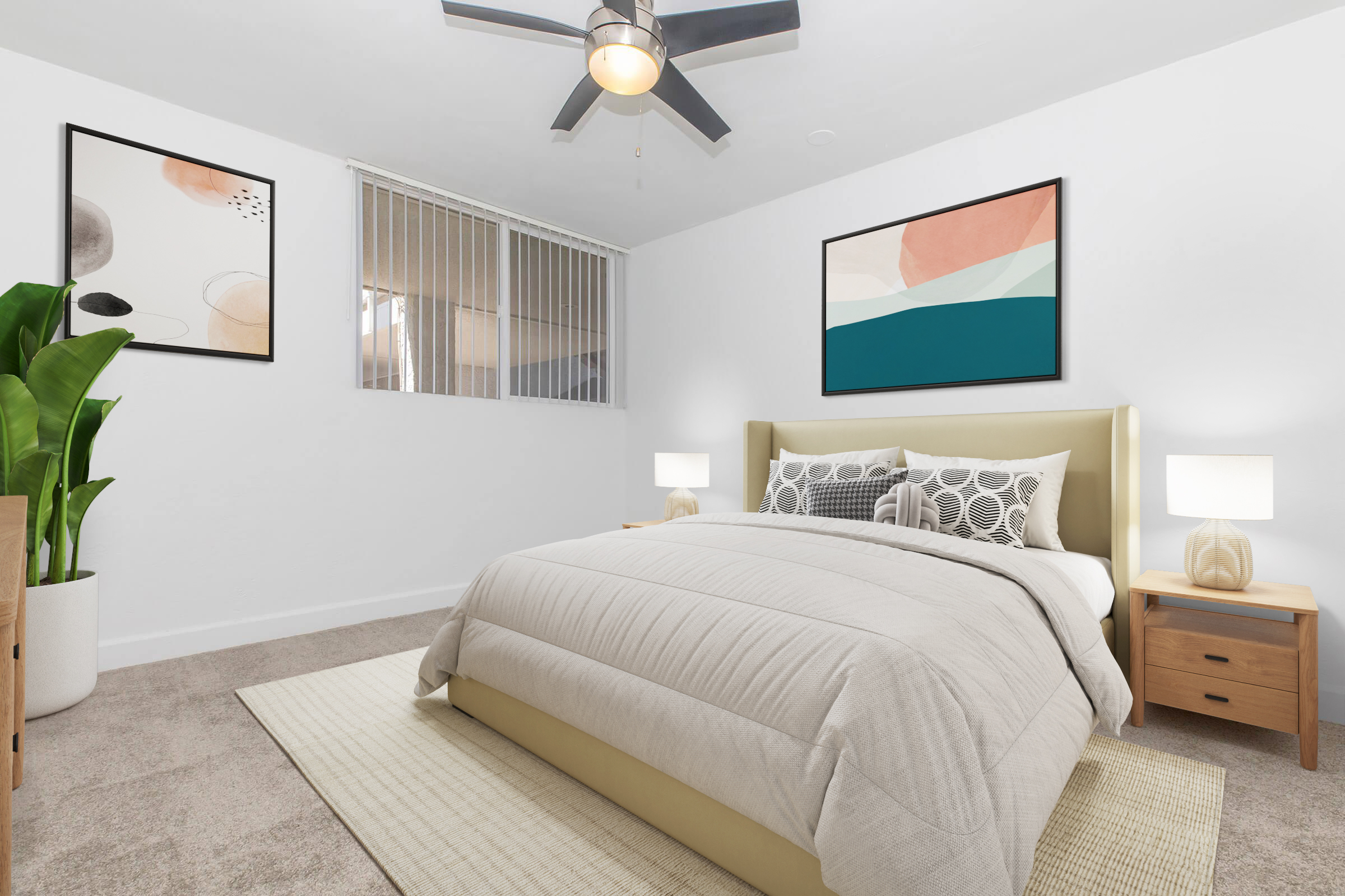 A cozy bedroom featuring a light-colored bed with a neatly arranged quilt, two bedside tables with lamps, and artwork on the walls. A large green plant adds a touch of nature. The room has soft beige carpeting and a ceiling fan, creating a warm and inviting atmosphere.