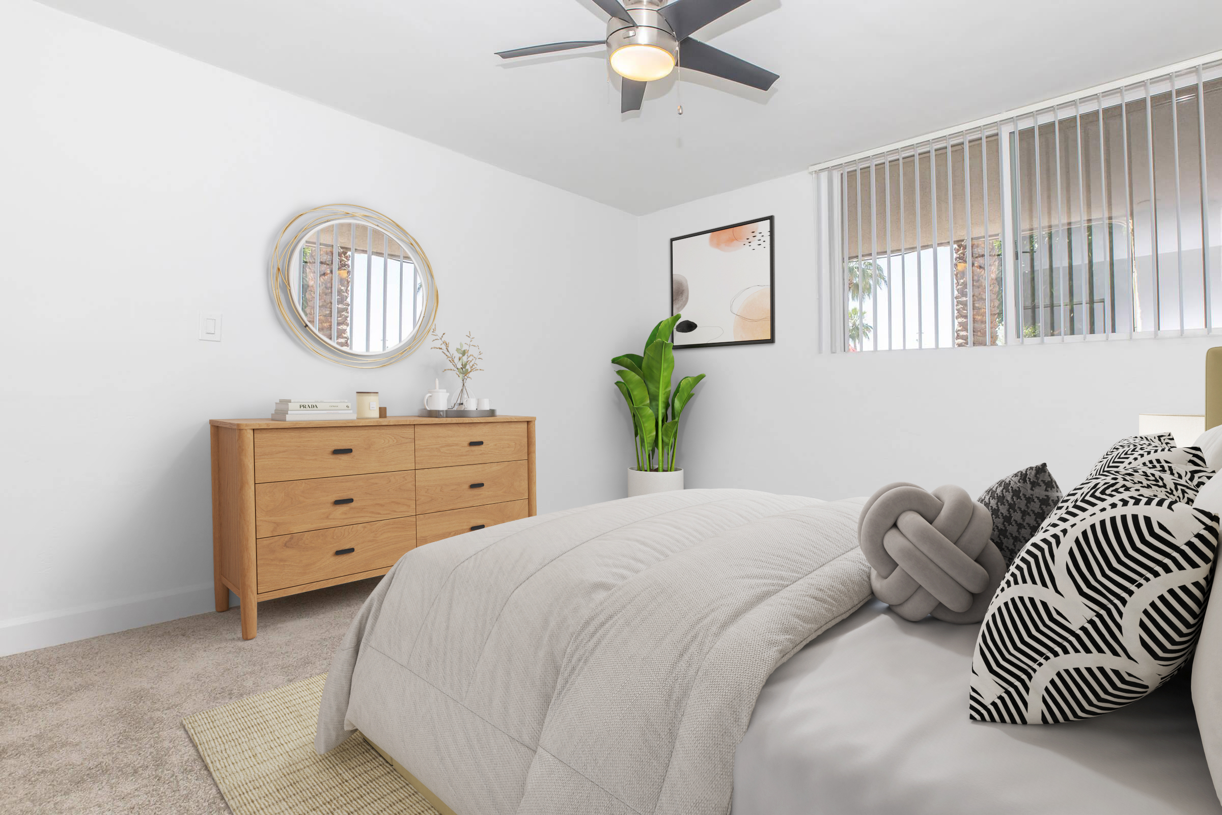 A well-lit bedroom featuring a cozy bed with decorative pillows, a stylish wooden dresser, a round mirror, and a potted plant. The walls are painted white, and there is a framed artwork on display. Large windows allow natural light to fill the space, creating a fresh and inviting atmosphere.