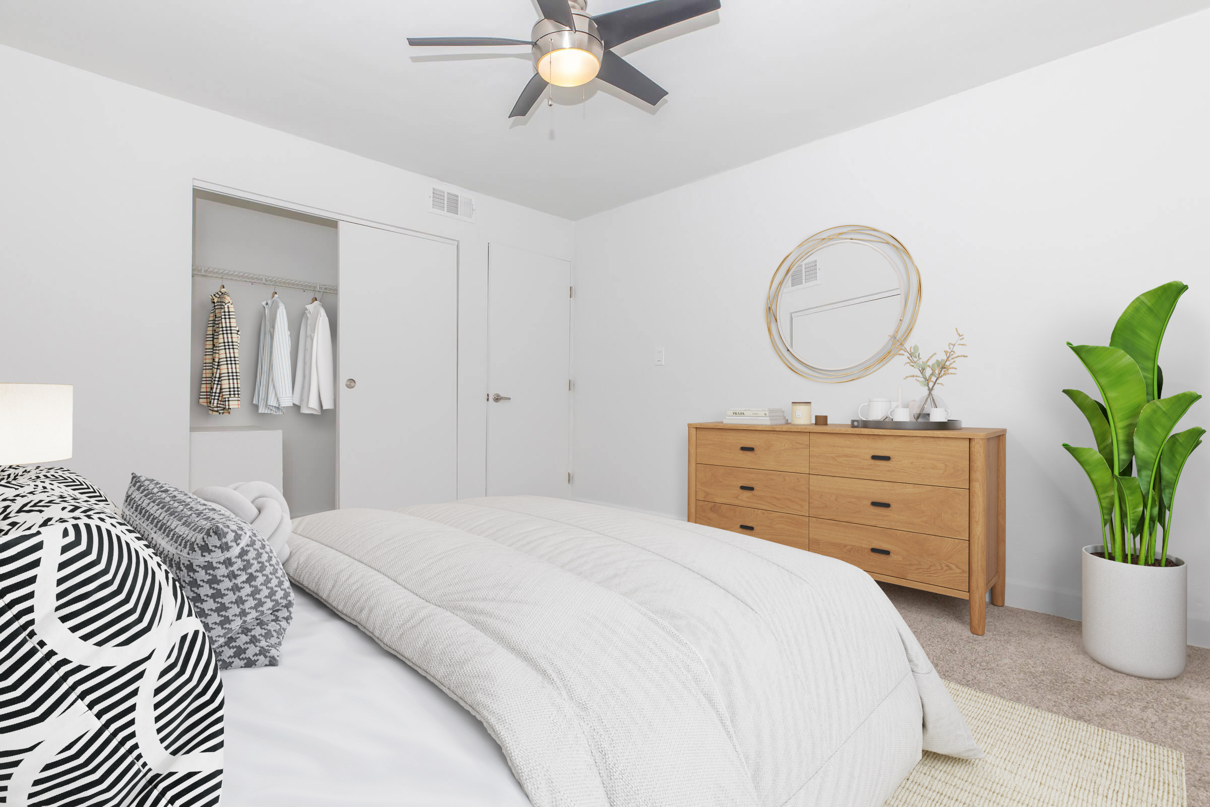 A modern bedroom featuring a bed with a light gray comforter and decorative pillows. There's a wooden dresser with multiple drawers, a round mirror above it, and a potted plant nearby. A closet door is slightly open, revealing hanging clothes, and a ceiling fan is visible above. The walls are painted white, creating a bright atmosphere.