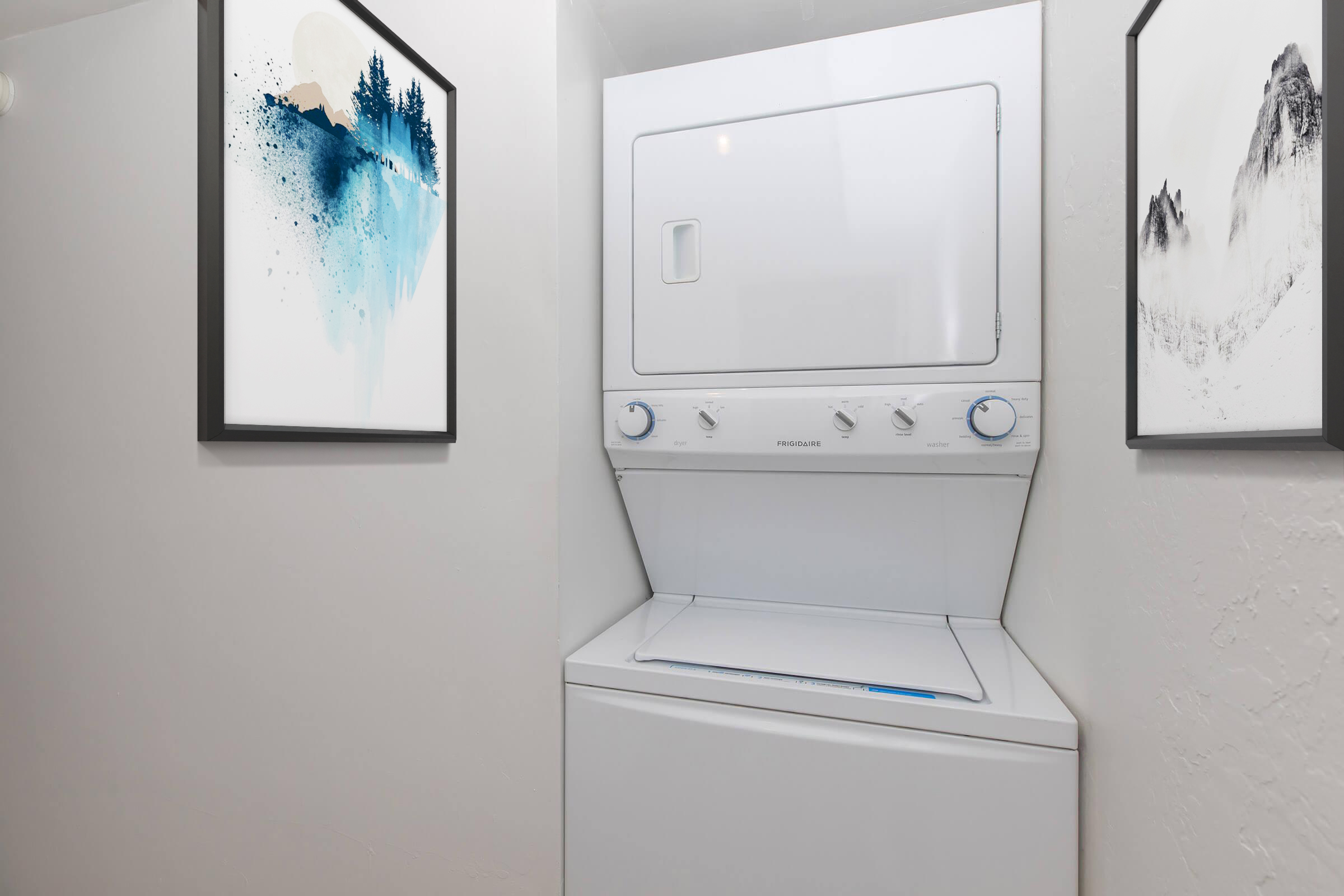 A stacked washer and dryer unit in a small, tidy laundry space. The appliances are white and placed between two framed abstract artworks featuring blue and gray tones on a light-colored wall.