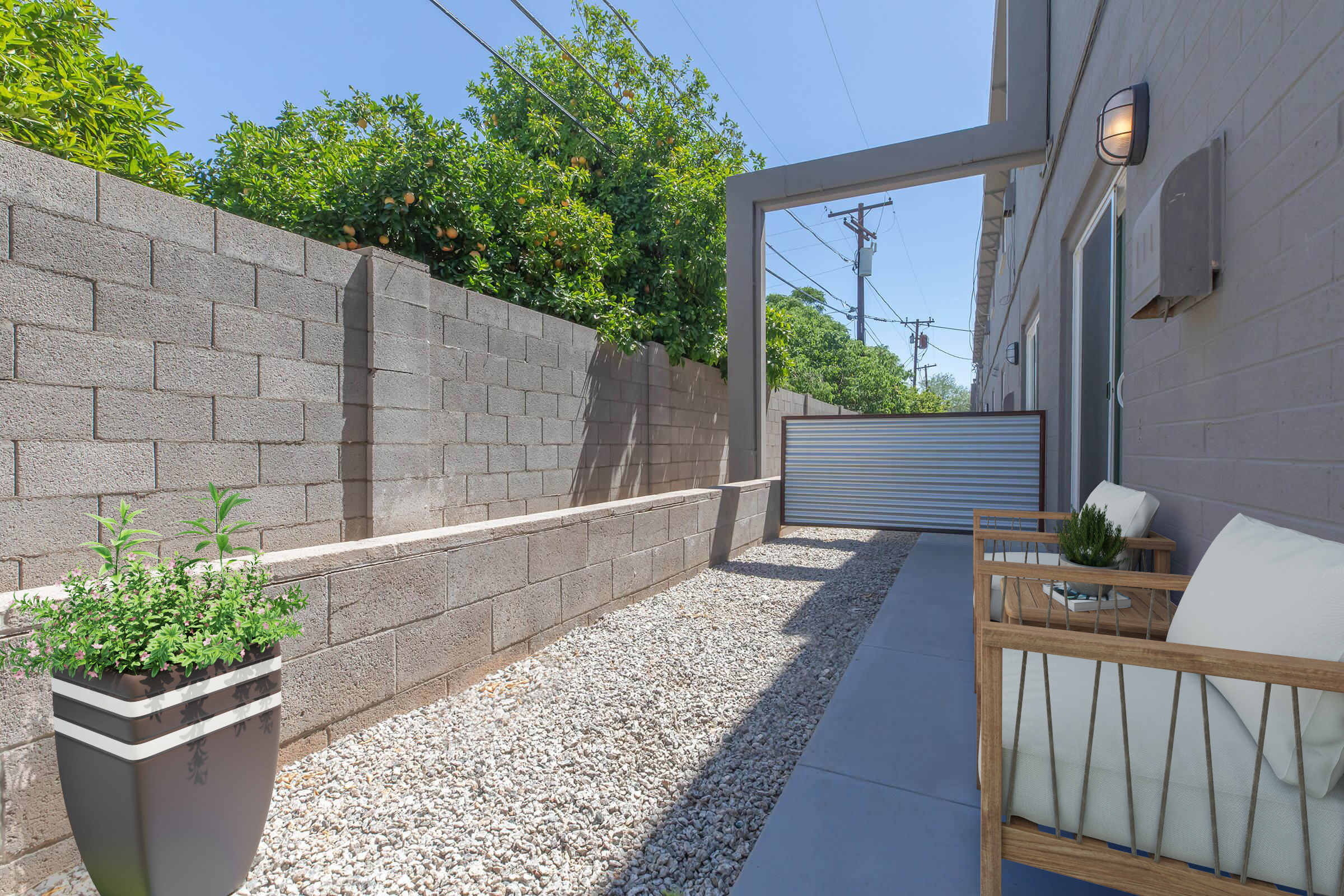 A pathway bordered by a stone wall, featuring a gravel surface and potted plants. On one side, there's a seating area with a wooden chair and cushions. In the background, a gate is visible along with greenery, under a bright blue sky.