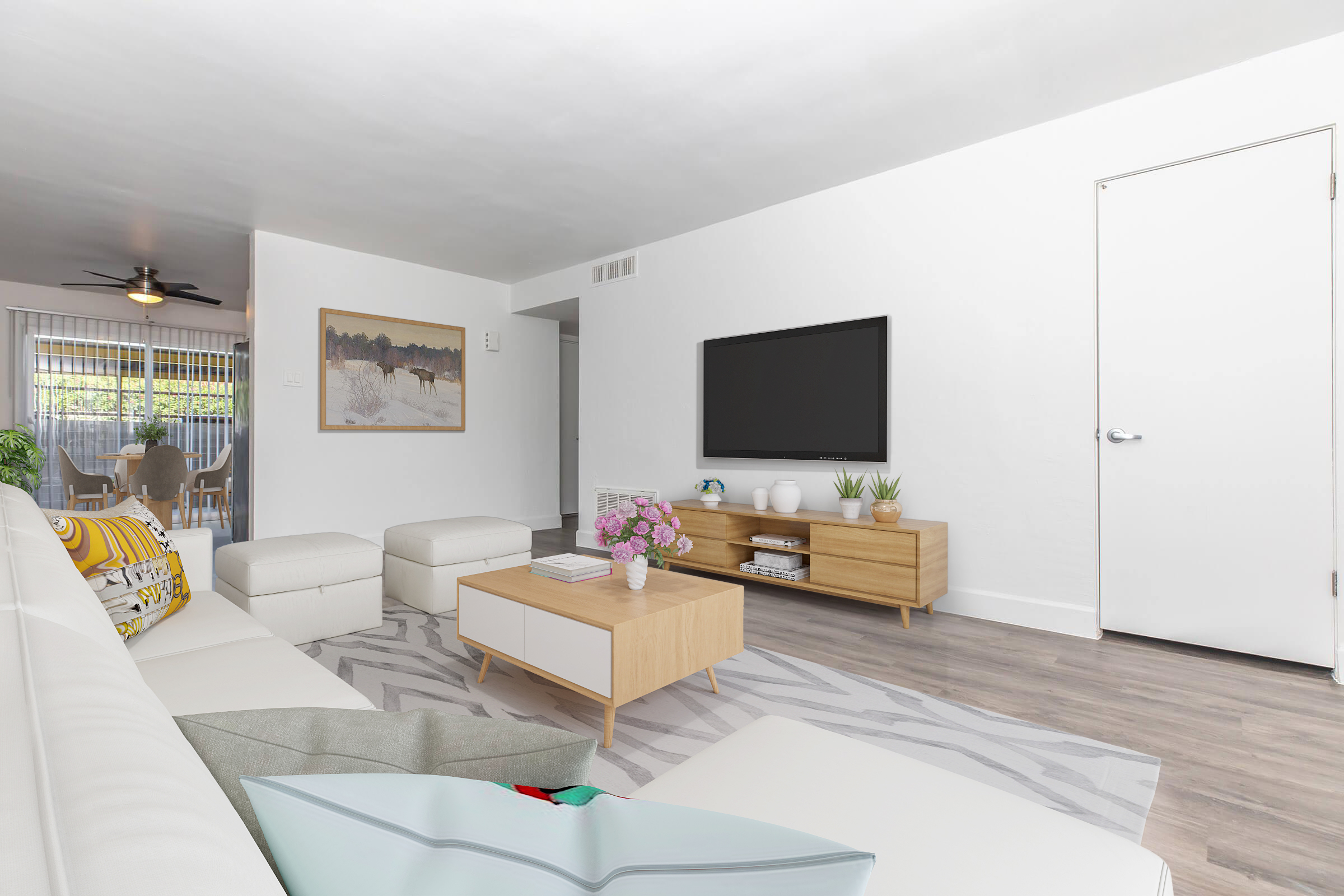 Bright and modern living room featuring a white sectional sofa, a light wood coffee table, and a television mounted on the wall. Decorative plants and a framed artwork add character. The room has light-colored walls, a ceiling fan, and large windows leading to an outdoor space.