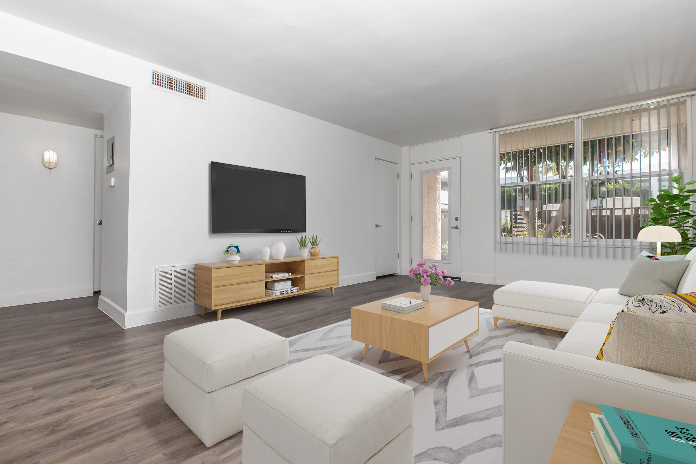 Bright and modern living room featuring a comfortable white sofa and two white ottomans. A wooden coffee table is in the center, adorned with a small plant and purple flowers. There is a large flat-screen TV mounted on the wall across from the seating. Natural light streams in from a door leading outside, enhancing the airy feel of the space.
