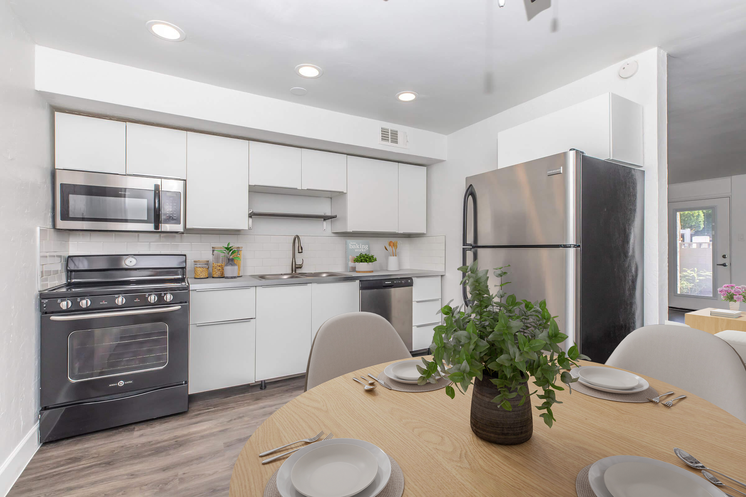 A modern kitchen featuring white cabinets, stainless steel appliances including a refrigerator, oven, and microwave. A dining table with four chairs is set for a meal, complete with plates and a small green plant centerpiece. Natural light comes from a window, enhancing the inviting atmosphere.