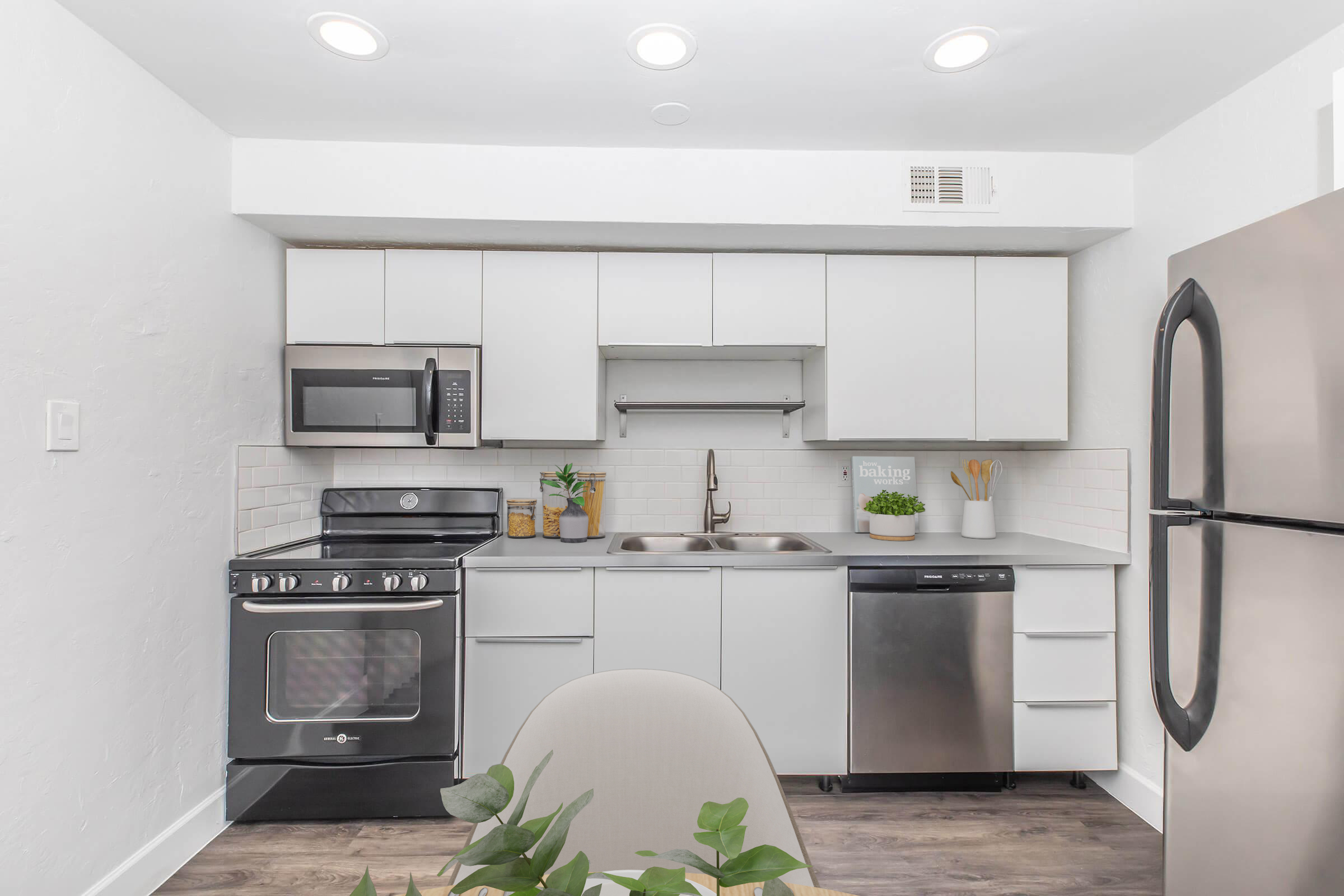 A modern kitchen featuring white cabinets, stainless steel appliances including an oven, microwave, and dishwasher. The sink is centrally located with a minimalist design. The countertops are clean, and there are potted plants for decoration. Bright LED lighting illuminates the space, creating a clean and inviting atmosphere.