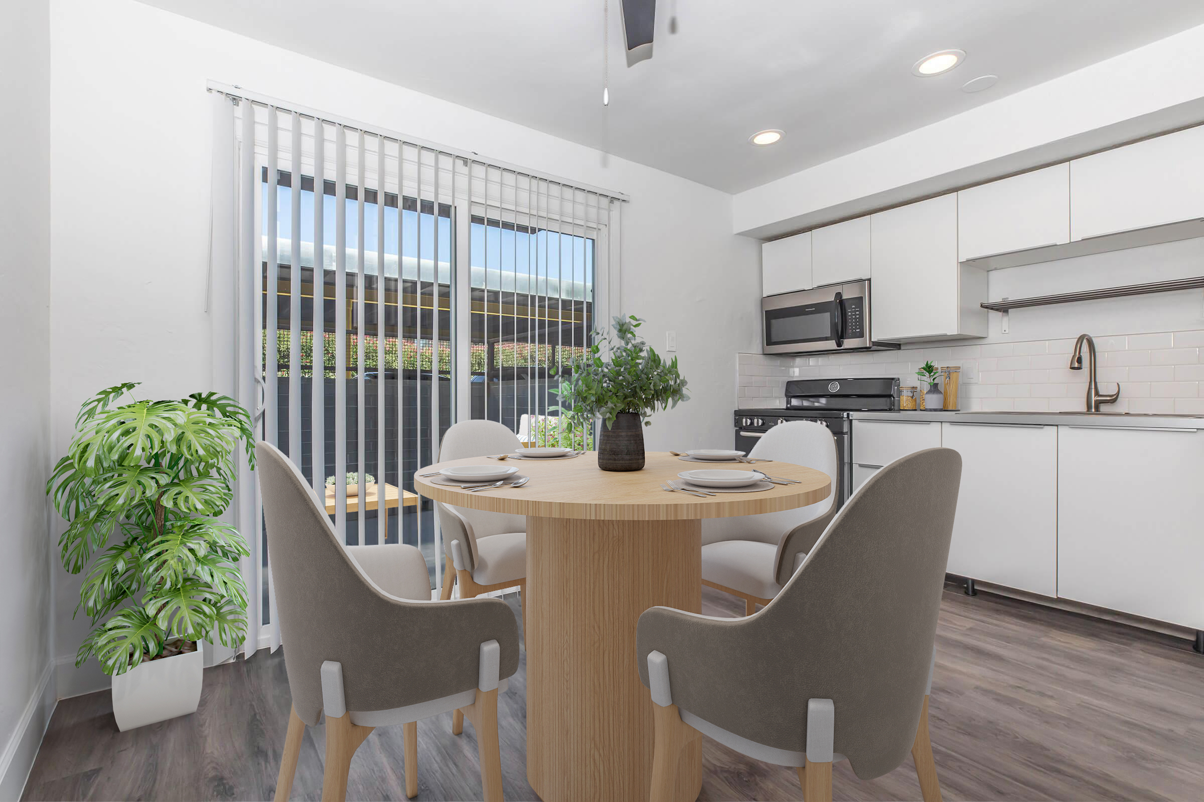 A modern dining area featuring a round wooden table surrounded by four gray chairs. There is a potted green plant in the corner and a large window with blinds providing natural light. The kitchen area includes sleek white cabinetry, a stainless steel microwave, and a stove. The floor is hardwood.