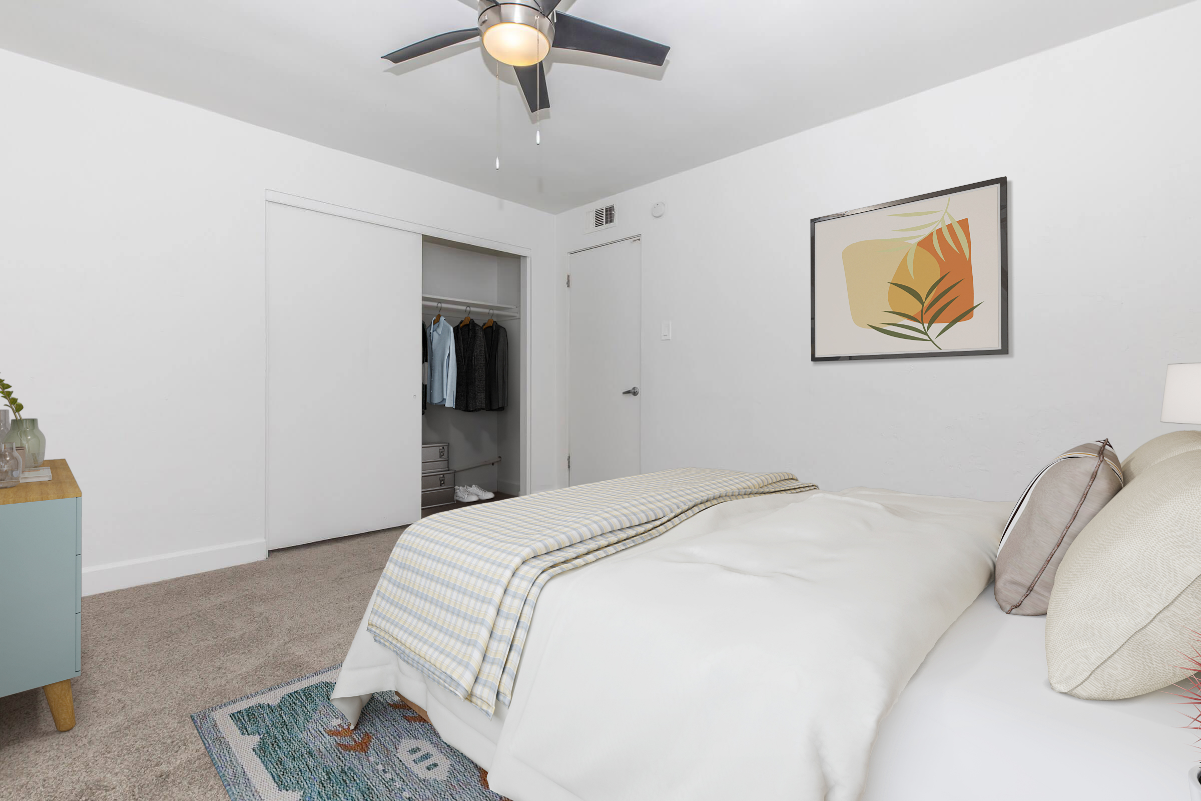 A cozy bedroom featuring a neatly made bed with a light-colored blanket, a small bedside table with a lamp, and a stylish wall art piece. There is a ceiling fan and a closet visible in the background, which contains neatly hung clothes. The room has a minimalistic, bright aesthetic with neutral tones.