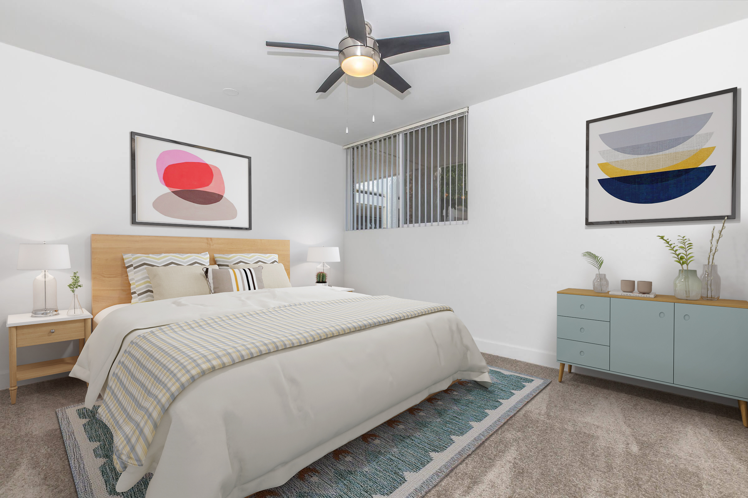 A modern bedroom featuring a wooden bed frame with light bedding, decorative pillows, and two bedside tables with lamps. There are colorful abstract artworks on the walls, a ceiling fan, and a blue sideboard with decorative items. A window with blinds allows natural light to enter the space.