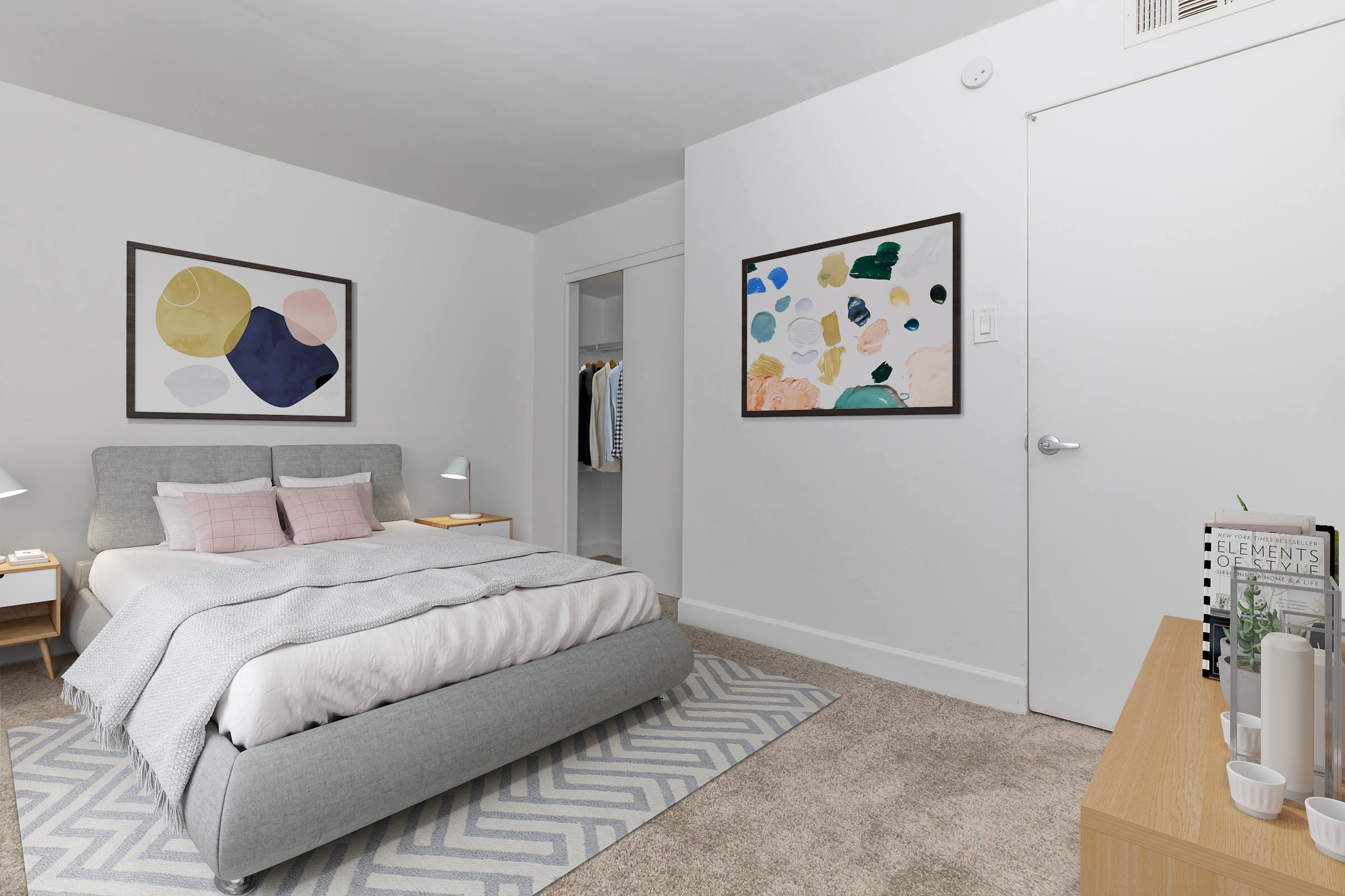 A cozy bedroom featuring a gray upholstered bed with light pink pillows, a patterned area rug, and two abstract art pieces on the walls. A wooden nightstand with lamps sits beside the bed, and there’s a closet with clothes visible in the background. A white door leads to another area of the room.
