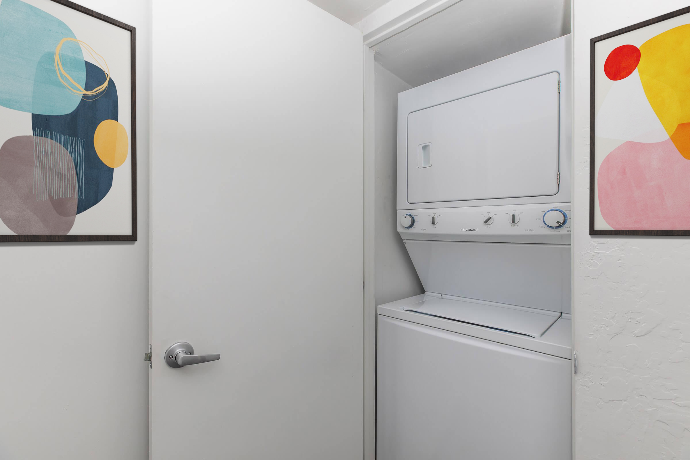 A small laundry area featuring a stacked washer and dryer, enclosed by white doors. On either side of the appliances, there are framed abstract art pieces with colorful shapes in shades of blue, yellow, and pink, adding a modern touch to the space.