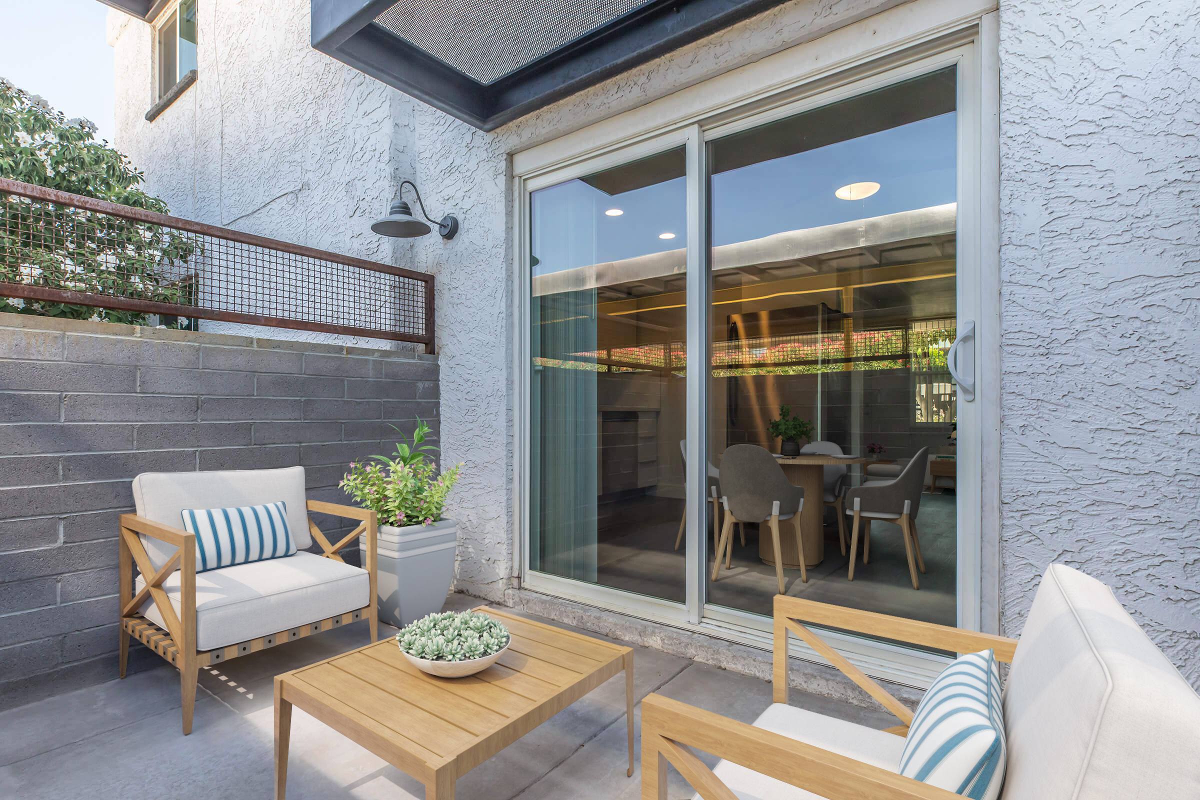 A cozy outdoor patio featuring two light-colored armchairs with striped cushions, a wooden coffee table with a small potted plant, and a view of a glass sliding door leading into a well-lit interior space. Surrounding walls are made of gray stone, creating a serene atmosphere.