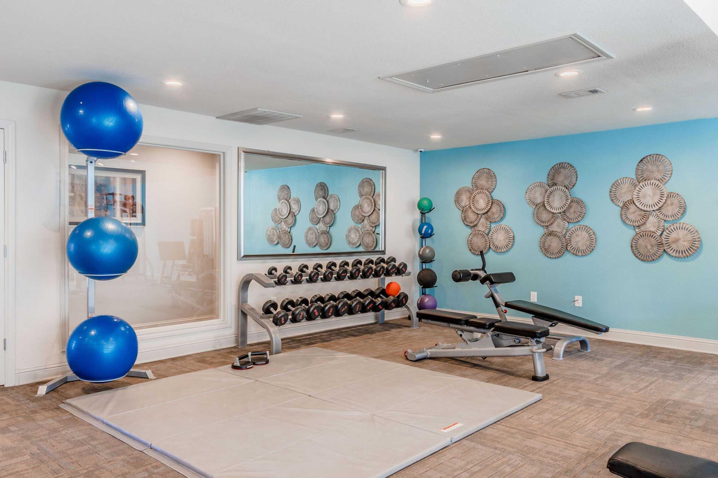 A modern gym interior featuring an array of exercise equipment. There are three large blue exercise balls, a weight bench, a collection of dumbbells on a rack, and a padded exercise mat on the floor. The walls are painted light blue, adorned with decorative wall art, and large mirrors reflect the space.