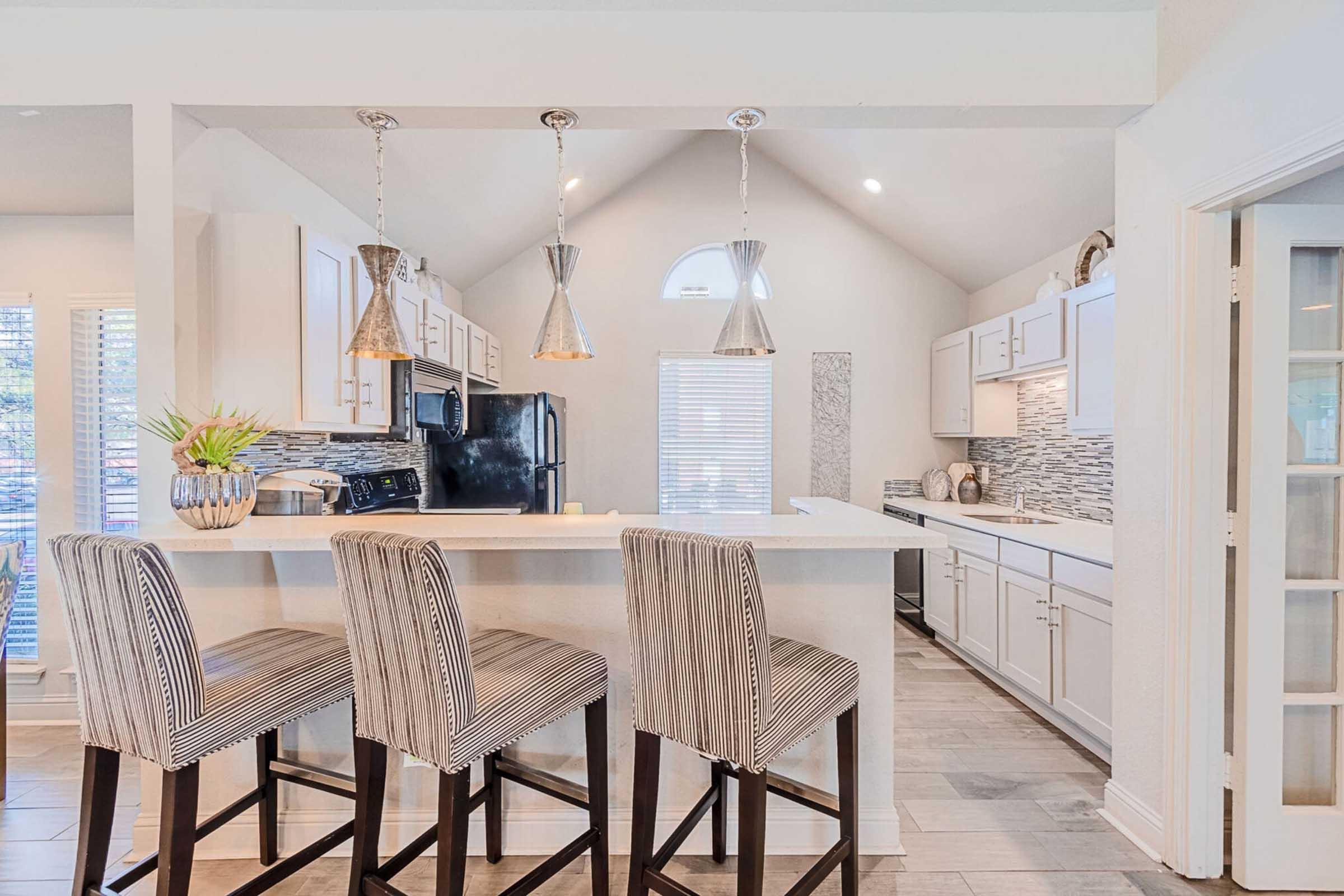 Bright and modern kitchen featuring a large island with three bar stools, pendant lighting, white cabinetry, and a stylish backsplash. Natural light streams in through windows, highlighting the spacious layout and contemporary appliances.