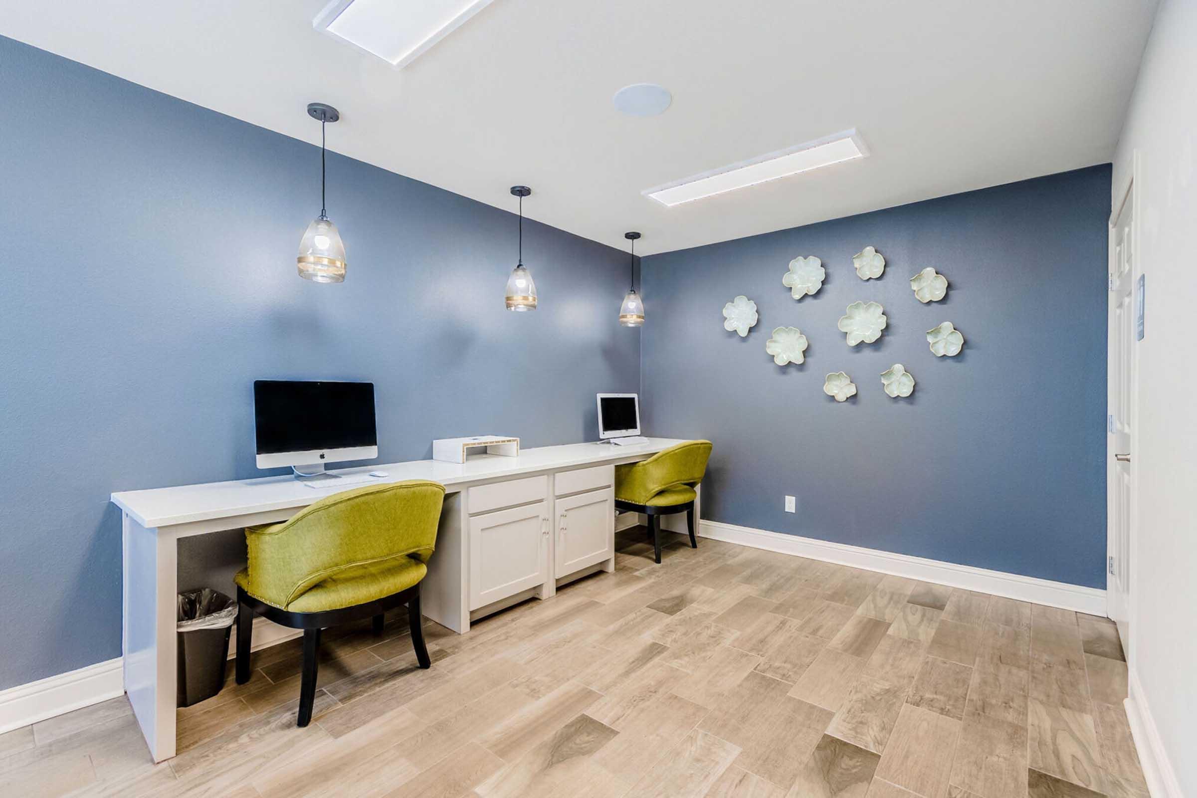 A modern workspace featuring two white desks with computers, stylish green chairs, and decorative wall accents. The walls are painted a rich blue, and pendant lights hang from the ceiling, creating a bright and inviting atmosphere. A wall sculpture of white flowers adds a touch of elegance.
