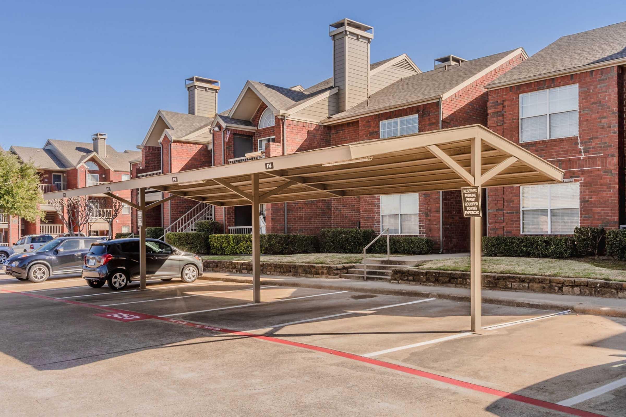 A row of covered parking spaces in front of brick apartment buildings. The area is well-maintained, with trimmed hedges and a clear sky. Several cars are parked under the canopy, and there are numbers indicating specific spots. The design features a modern, suburban aesthetic.