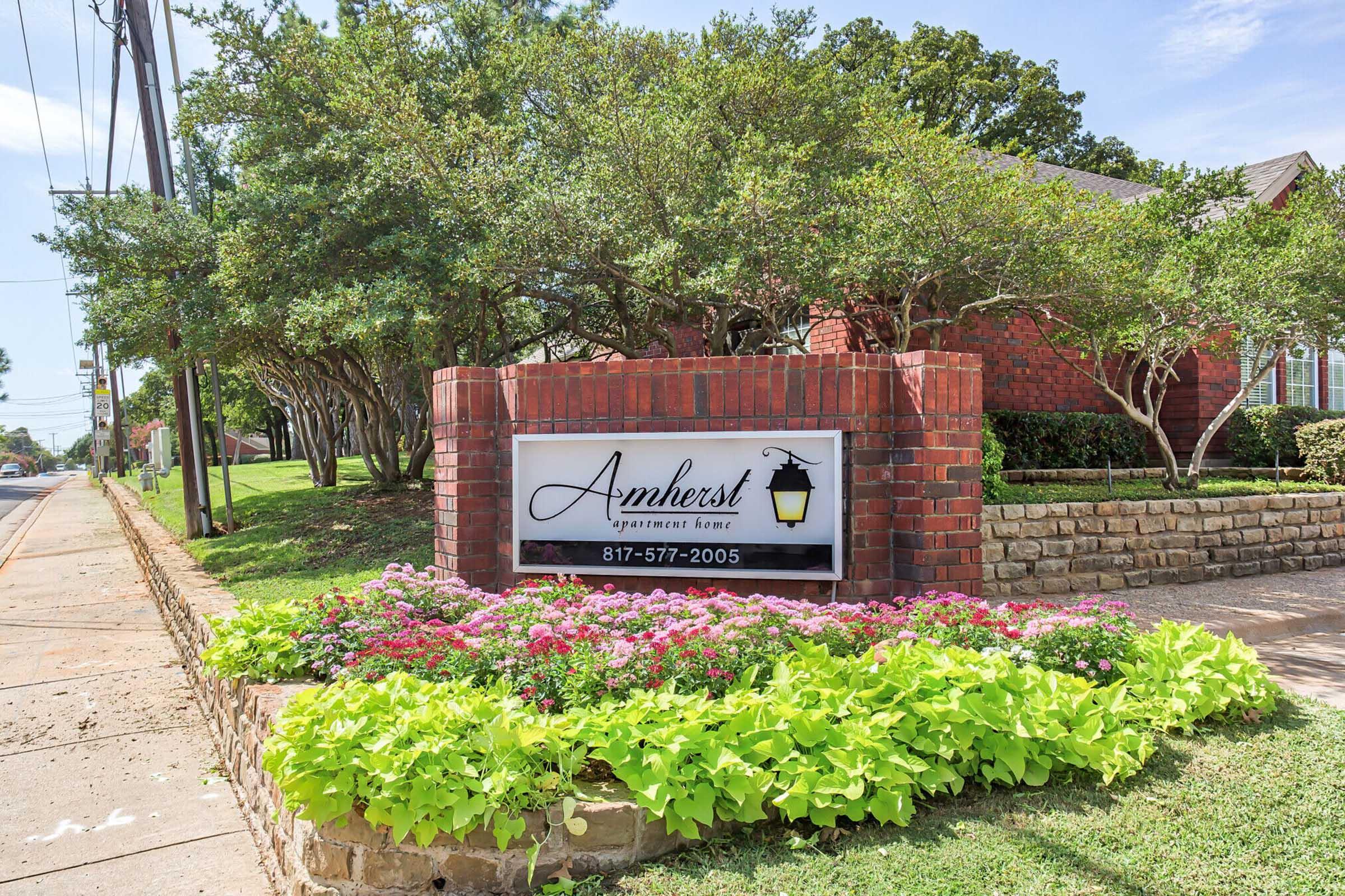 Sign for Amherst apartments, featuring the name prominently displayed with a decorative lamp icon. Surrounded by well-maintained landscaping, including colorful flowers and green bushes, with a residential area visible in the background. Clear blue sky overhead.