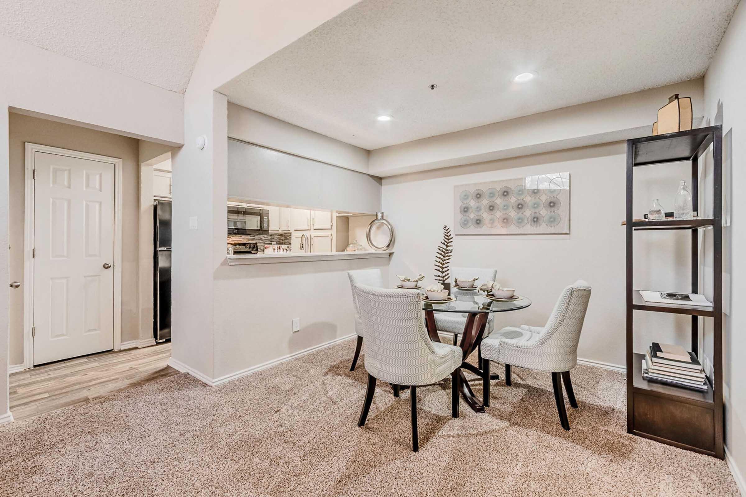 A cozy dining area featuring a round glass table set for four, surrounded by stylish light-colored chairs. The space is well-lit, with soft lighting and a textured ceiling. In the background, there's a kitchenette with modern appliances and a decorative shelf displaying a pinecone and books.