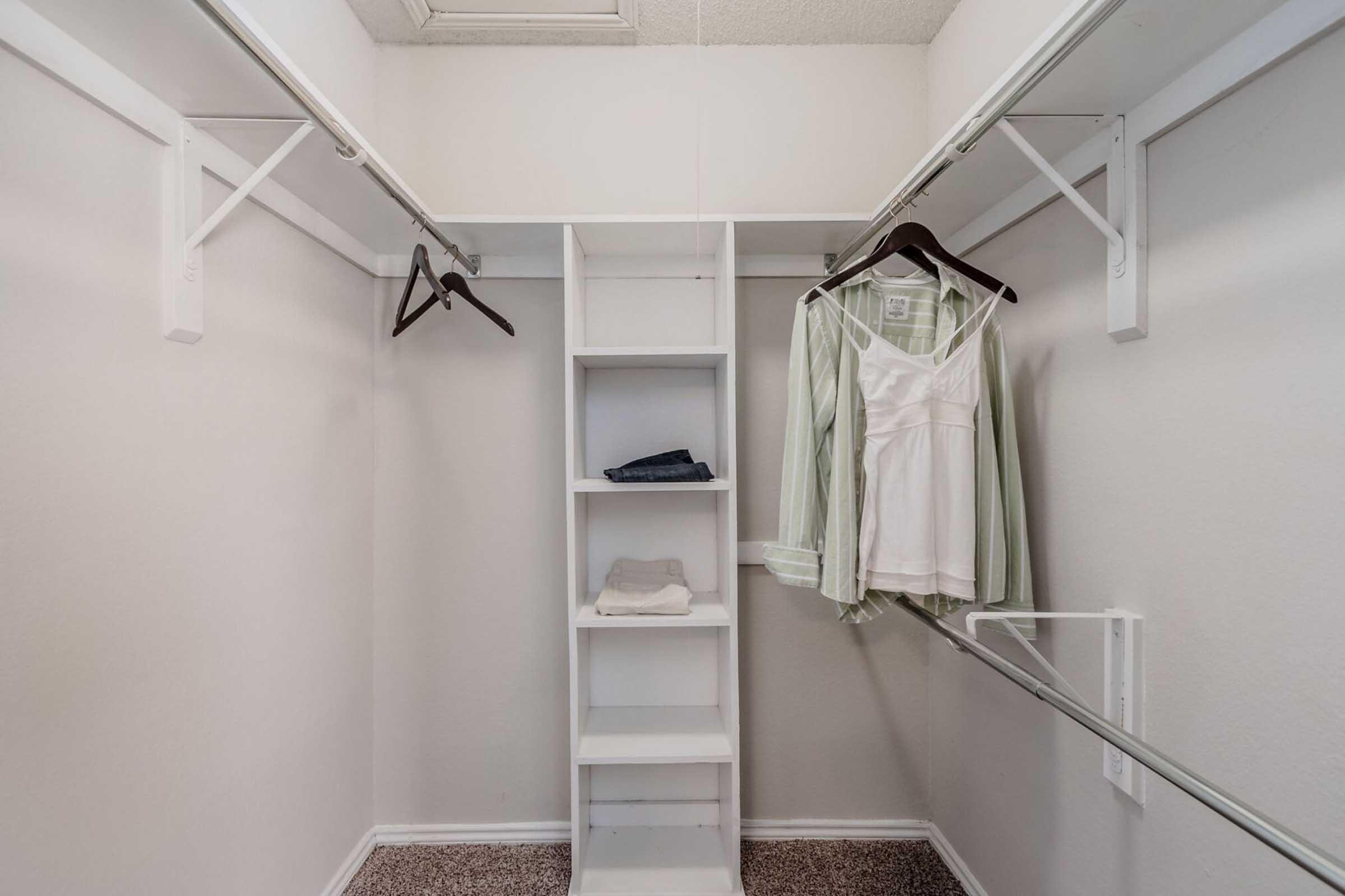 Empty closet with white walls and carpeted floor. There are two hanging rods with clothing on hangers—one shirt and some folded clothes on a shelf. The space is organized with a shelf unit in the center for additional storage.