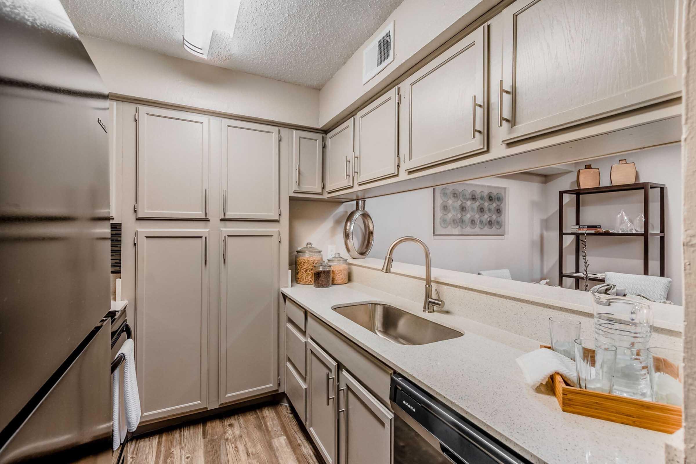 A modern kitchen featuring light gray cabinets, a stainless steel sink, and a countertop. The kitchen includes a black refrigerator, jars on the countertop, and a decorative shelf in the background. Soft lighting and a clean design create a welcoming atmosphere.