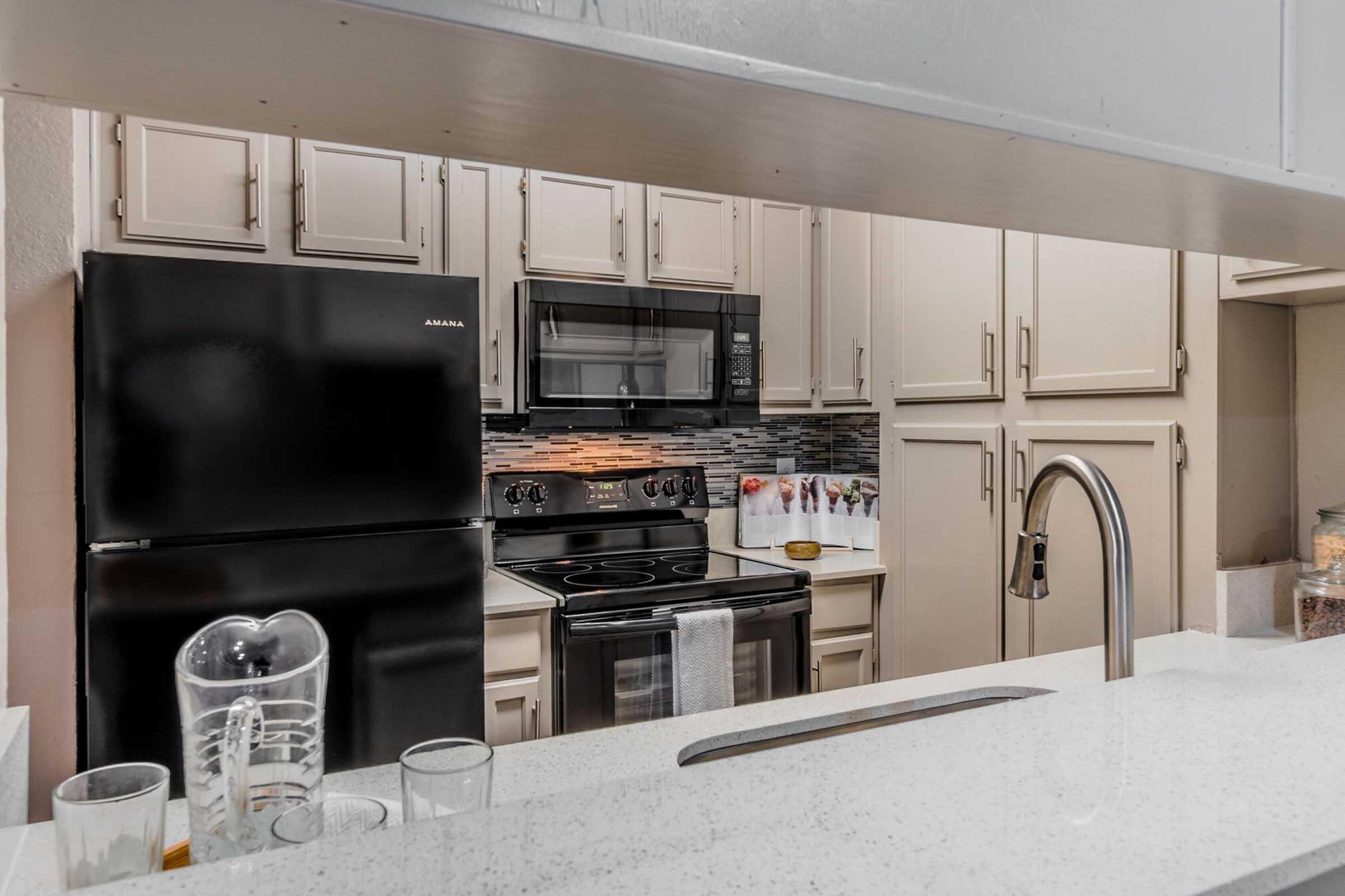 A modern kitchen featuring a black refrigerator and microwave, a silver stove, and beige cabinetry. The countertop is white with a sink and faucet visible. Decorative jars are placed near the sink, and there’s a backsplash with textured tiles. Glassware and various utensils are scattered across the countertop.