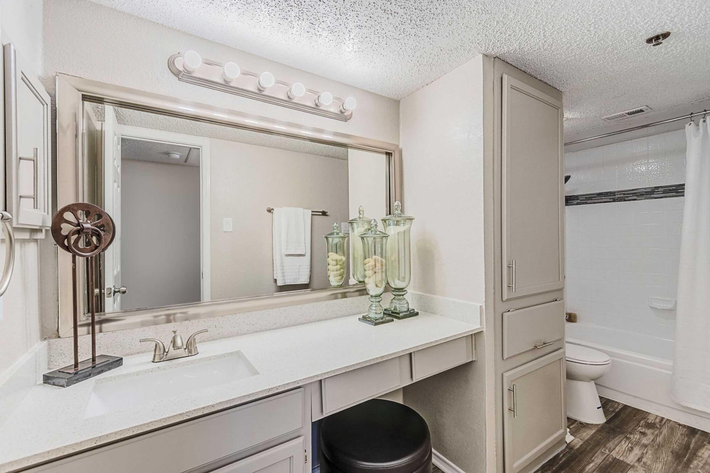 A modern bathroom featuring a double-sink vanity with a white countertop, a large mirror with overhead lighting, and decorative glass containers. There's a toilet and a shower area visible in the background, along with neutral-colored walls and towels hanging nearby.
