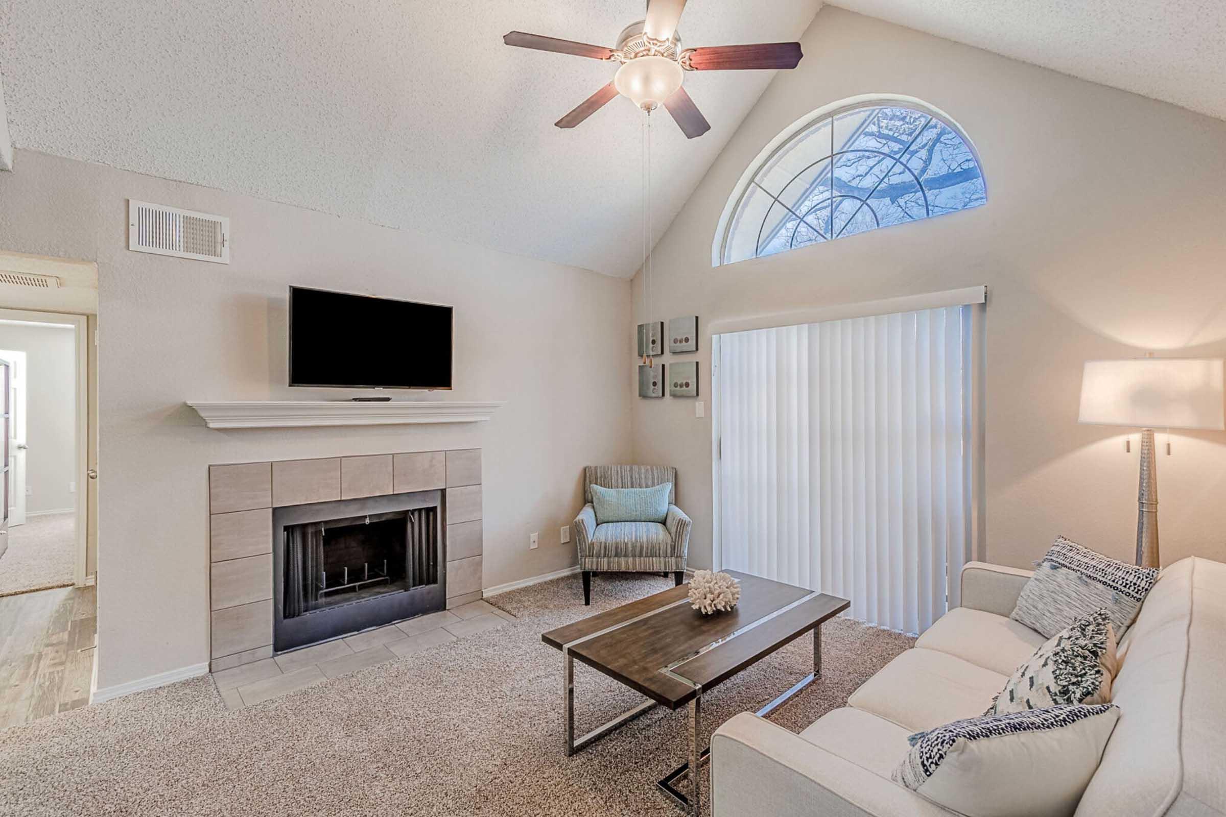 Bright living room featuring a white couch, a wooden coffee table, and a modern armchair. There's a fireplace with a TV mounted above it on the wall. A large arched window allows natural light to illuminate the space, and there are sliding glass doors covered with blinds, enhancing the cozy atmosphere.