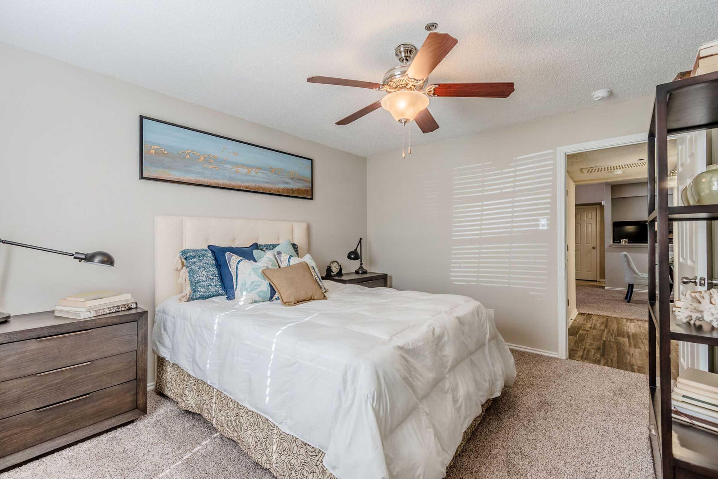 Brightly lit bedroom featuring a queen-sized bed with white bedding and decorative pillows. There is a nightstand with a lamp on one side and a dark wooden bookshelf on the other. A ceiling fan adds a cozy touch, and a tasteful piece of artwork hangs above the bed. The room has a warm, inviting atmosphere.