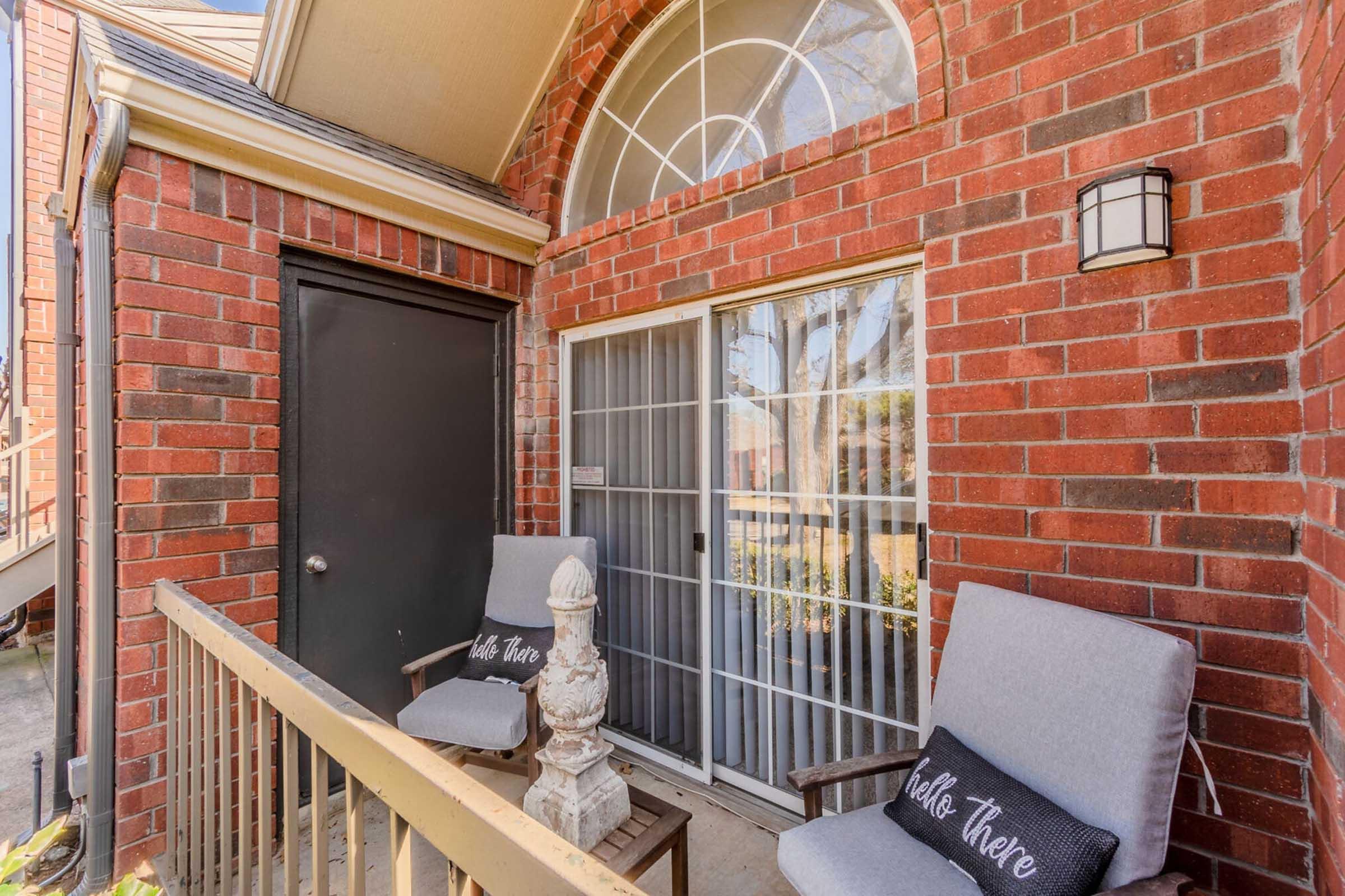 A cozy porch area featuring two gray chairs, one with a "Hello There" pillow, a small table, and a decorative stone accent. The entrance has a brick wall and a large arched window. A door leads into the home, and trees can be seen in the background. The setting is warm and inviting.