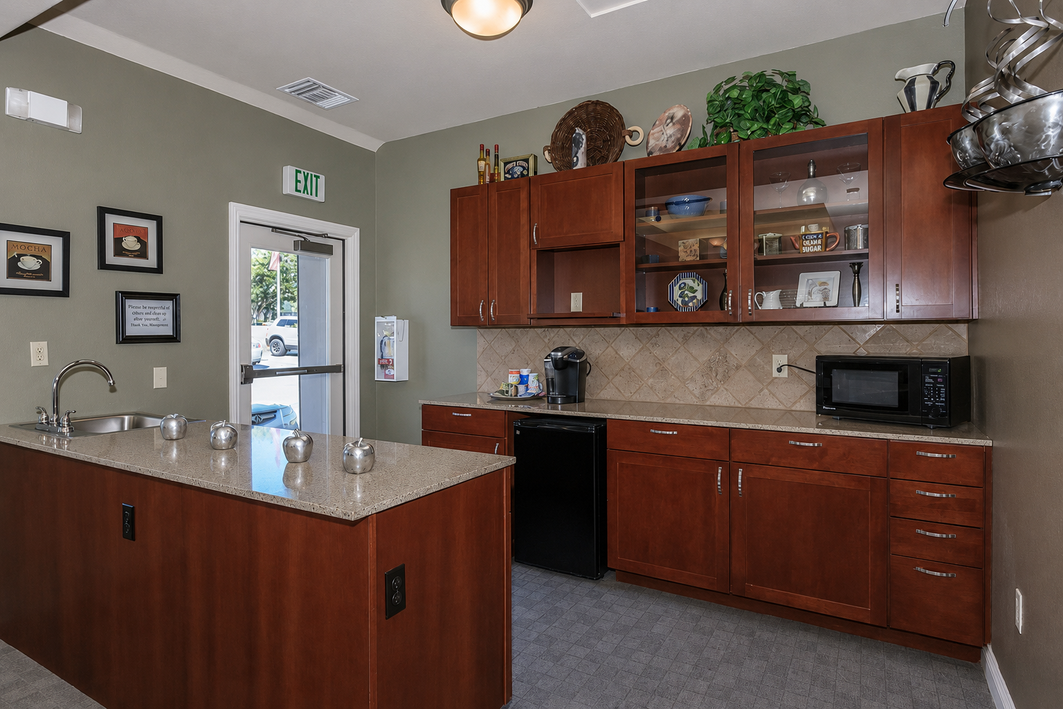 Modern kitchen with dark wood cabinetry, granite countertops, and a tiled backsplash. Features include a stainless steel sink, black refrigerator, microwave, coffee maker, and decorative plates. Natural light comes through a glass door leading outside, with a stylish overhead light fixture and green plants for accent.