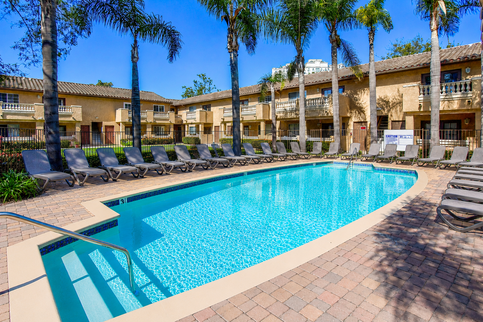 A sparkling blue swimming pool surrounded by palm trees and lounge chairs, located in a sunny courtyard with two-story buildings in the background. The poolside area features paved bricks and a clear sky overhead.