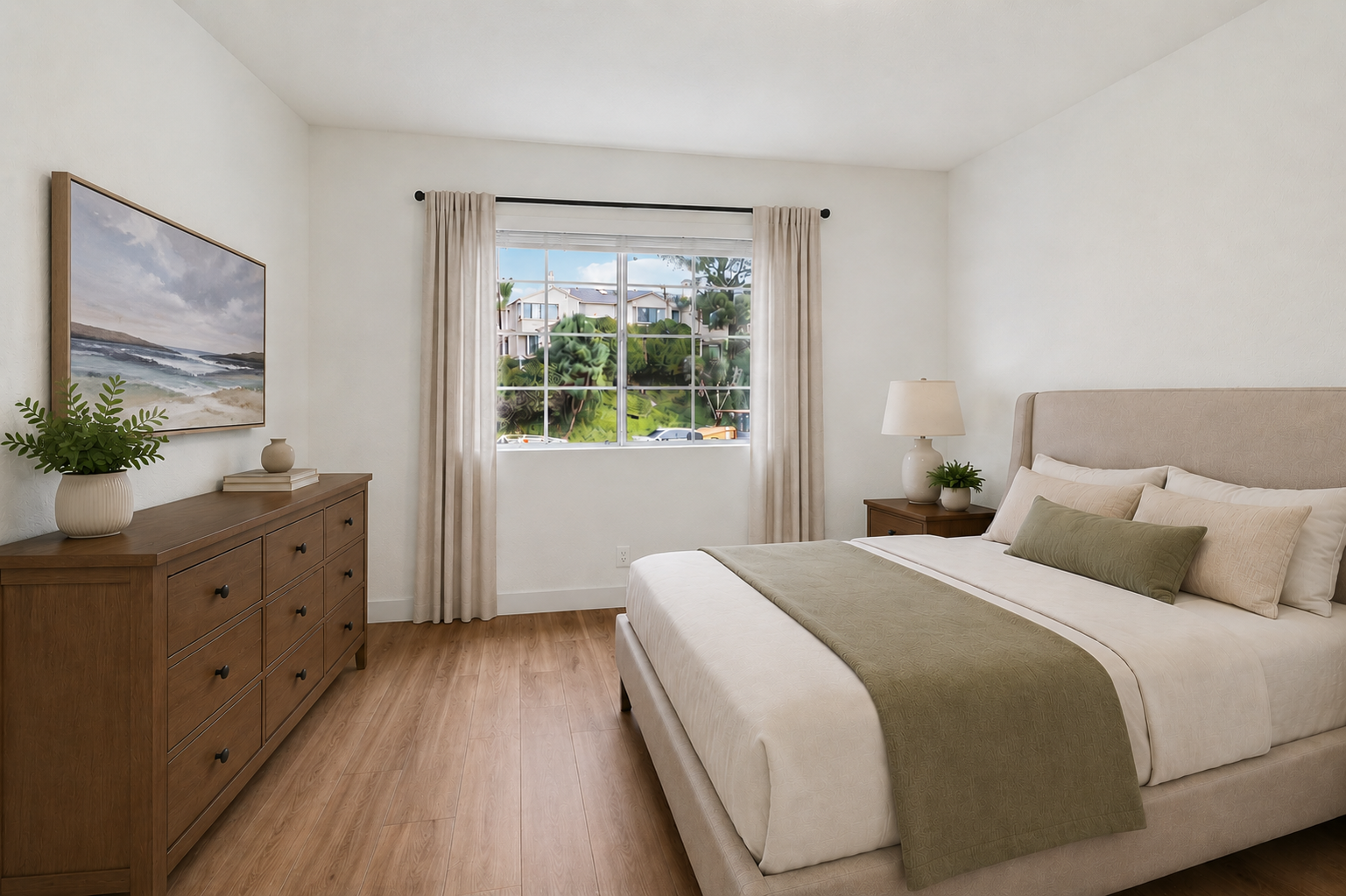A cozy bedroom featuring a neutral color palette. The room includes a large window with a view of greenery outside, a light-colored bed with decorative pillows, a bedside lamp, and a wooden dresser. Artwork depicting a beach scene hangs on the wall, adding a touch of tranquility to the space.