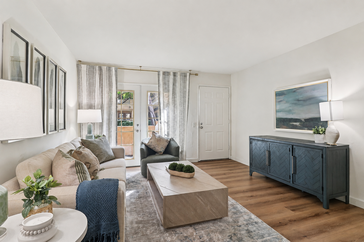 A cozy living room featuring a light-colored sofa adorned with decorative pillows, a sleek coffee table, and an elegant sideboard. Natural light streams in through glass doors leading to an outdoor space. The room is decorated with contemporary artwork, indoor plants, and soft lighting, creating a welcoming atmosphere.