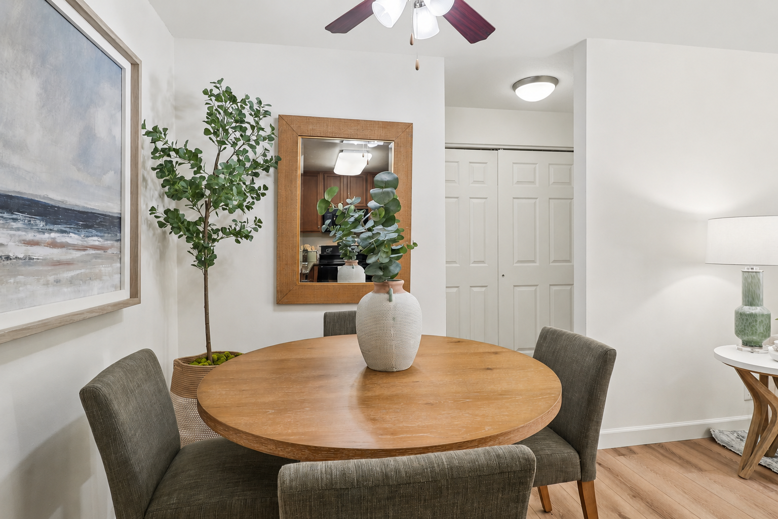 A cozy dining area featuring a round wooden table with a vase of greenery at its center. Surrounding the table are four upholstered chairs. A large framed painting of a coastal scene hangs on the wall, alongside a mirror that reflects the room. A light fixture and a cupboard are visible in the background.