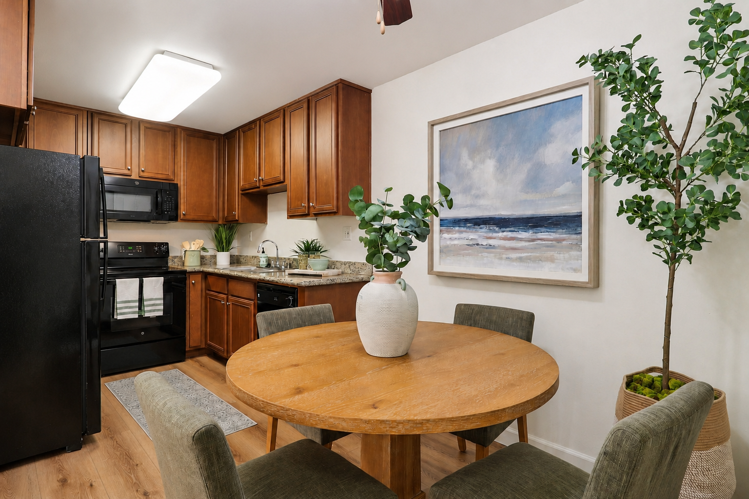 A cozy kitchen featuring dark wooden cabinetry, black appliances, and a circular wooden dining table surrounded by green upholstered chairs. A large painting of a seascape hangs on the wall, and a potted plant adds a fresh touch to the space. Warm lighting enhances the inviting atmosphere.