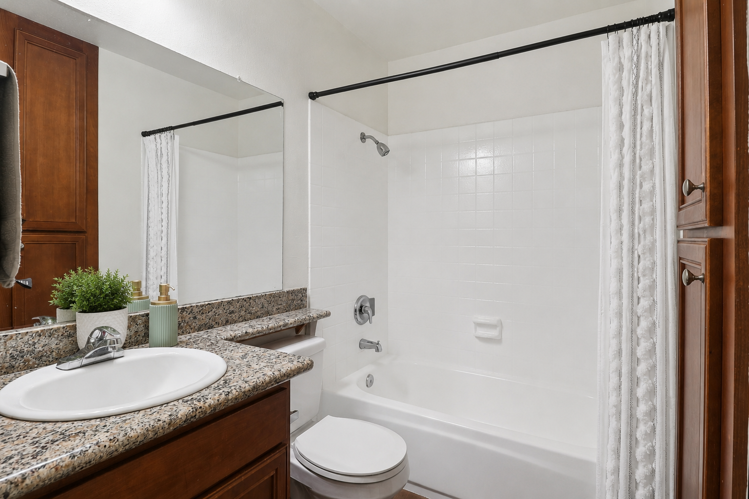 A clean, modern bathroom featuring a bathtub with a shower curtain, a sleek granite countertop with a small plant, and wooden cabinetry. The space is well-lit with a mirror above the sink and neutral-colored walls, creating a bright and welcoming atmosphere.