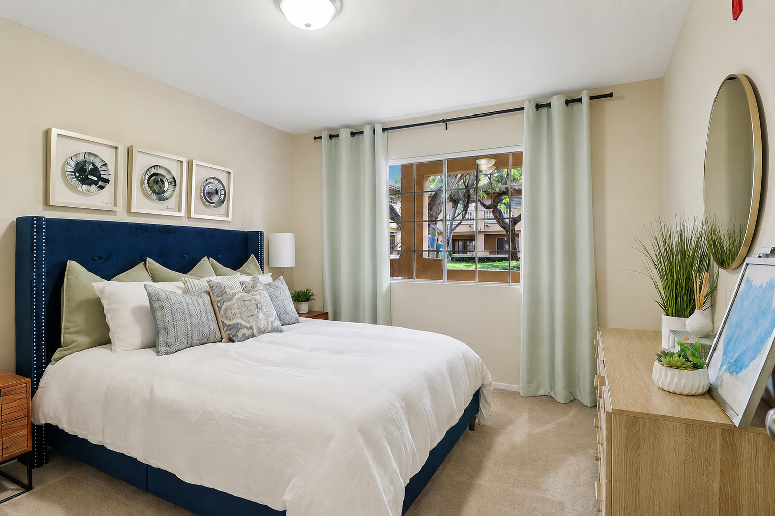 A cozy bedroom featuring a blue upholstered headboard with decorative pillows, a light-colored quilted duvet, and two bedside tables. There's a window with sheer curtains allowing natural light, framed artwork on the walls, and a dresser with plants and decorative items. The overall decor is modern and inviting.