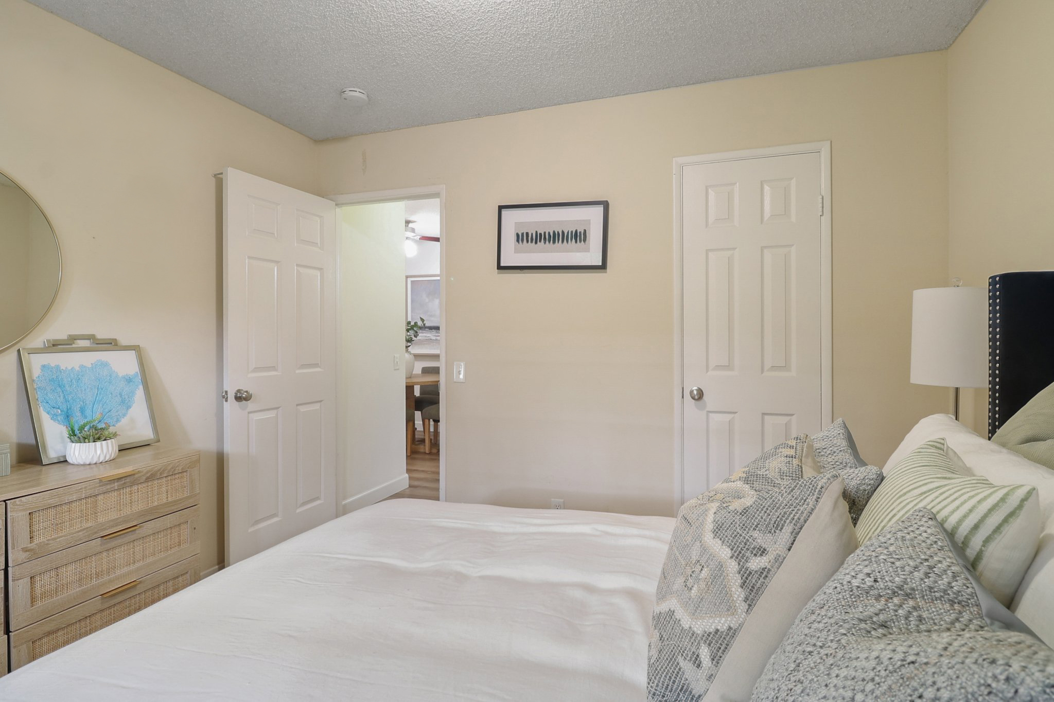 A cozy bedroom featuring a neatly made bed with decorative pillows, a wooden dresser with a blue abstract artwork, and two closed doors leading to other rooms. Natural light fills the space, creating a warm atmosphere. The walls are softly painted in beige, complementing the overall decor.