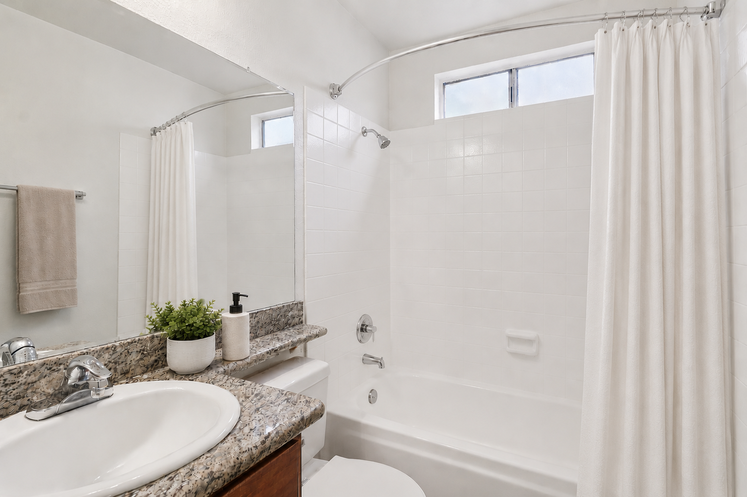 A clean and modern bathroom featuring a white bathtub with a shower curtain, a granite countertop with a sink, and a small potted plant. The walls are tiled in white, and there is a towel hanging on the rack. Natural light comes from a window above the bathtub.