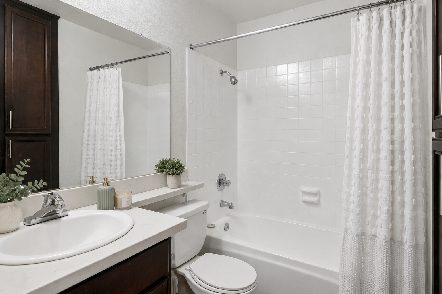 A clean and modern bathroom featuring a white bathtub with a shower curtain, a large mirror above a sink with a countertop, dark wooden cabinetry, and a small potted plant on the counter. The walls are painted a light color, creating a bright and inviting atmosphere.