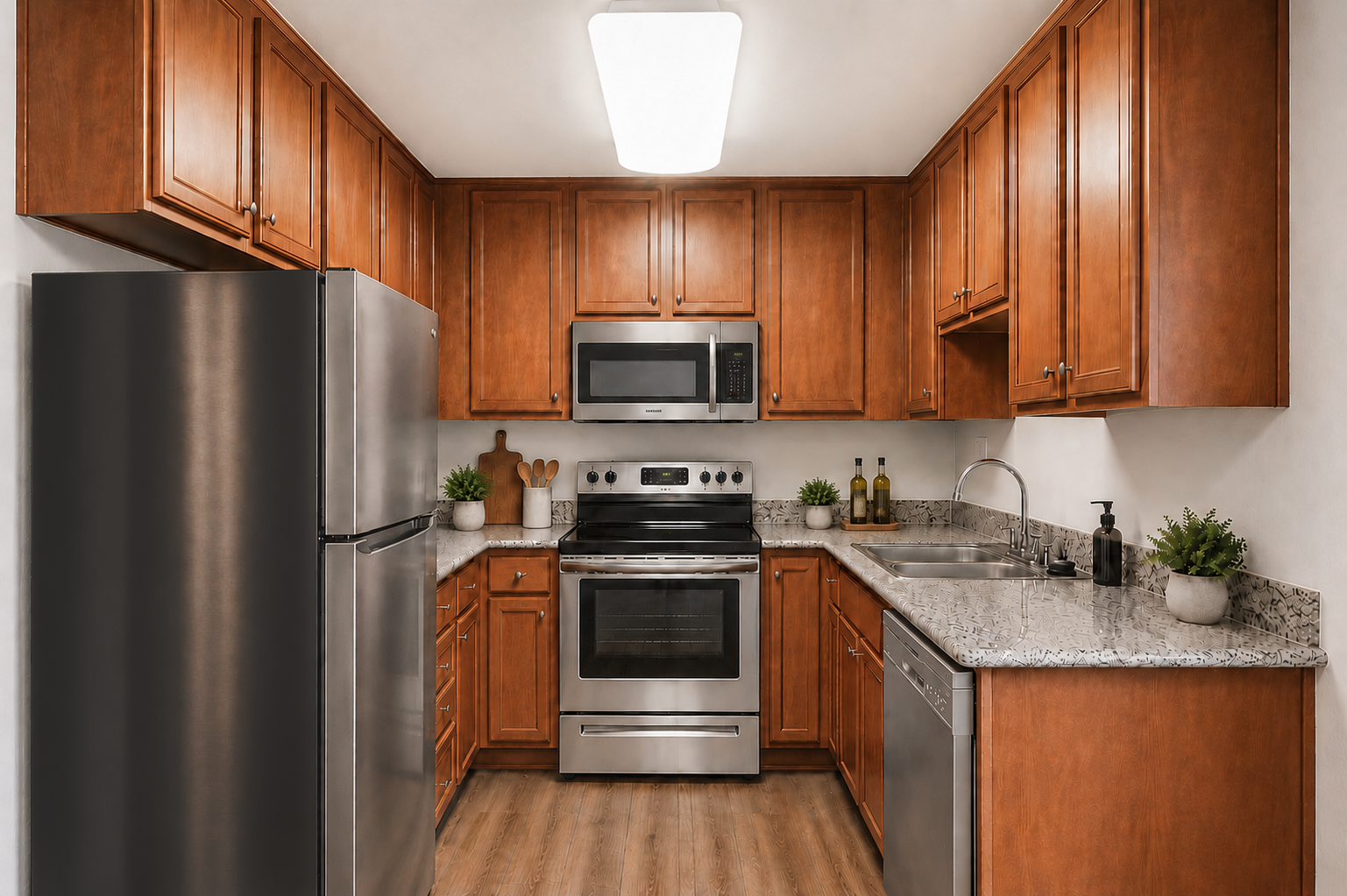 A modern kitchen featuring wooden cabinets, granite countertops, stainless steel appliances including a fridge, oven, and dishwasher, and a sink. There are decorative plants on the countertop and a wooden cutting board on the side, illuminated by overhead lighting.